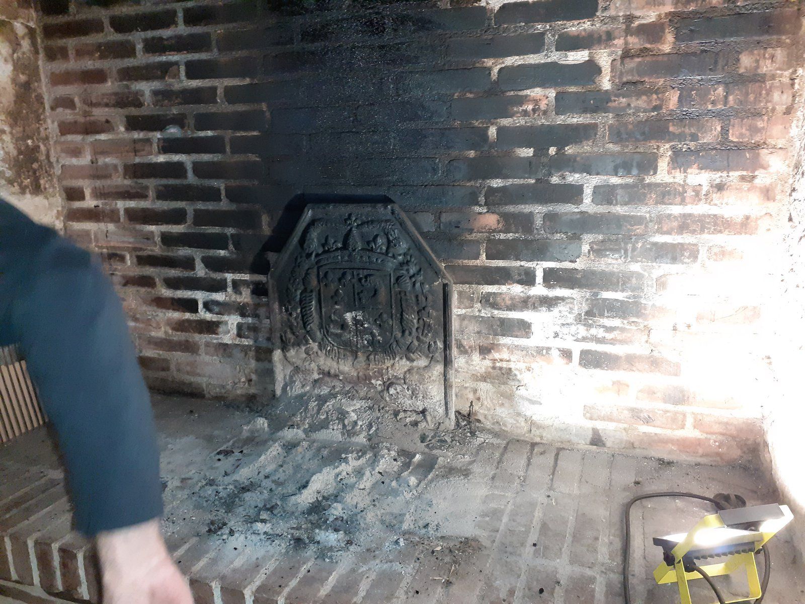 Cheminée en briques brûlées avec cendres et grille à feu ; un bras humain y entre par la gauche et un outil jaune se trouve à droite.