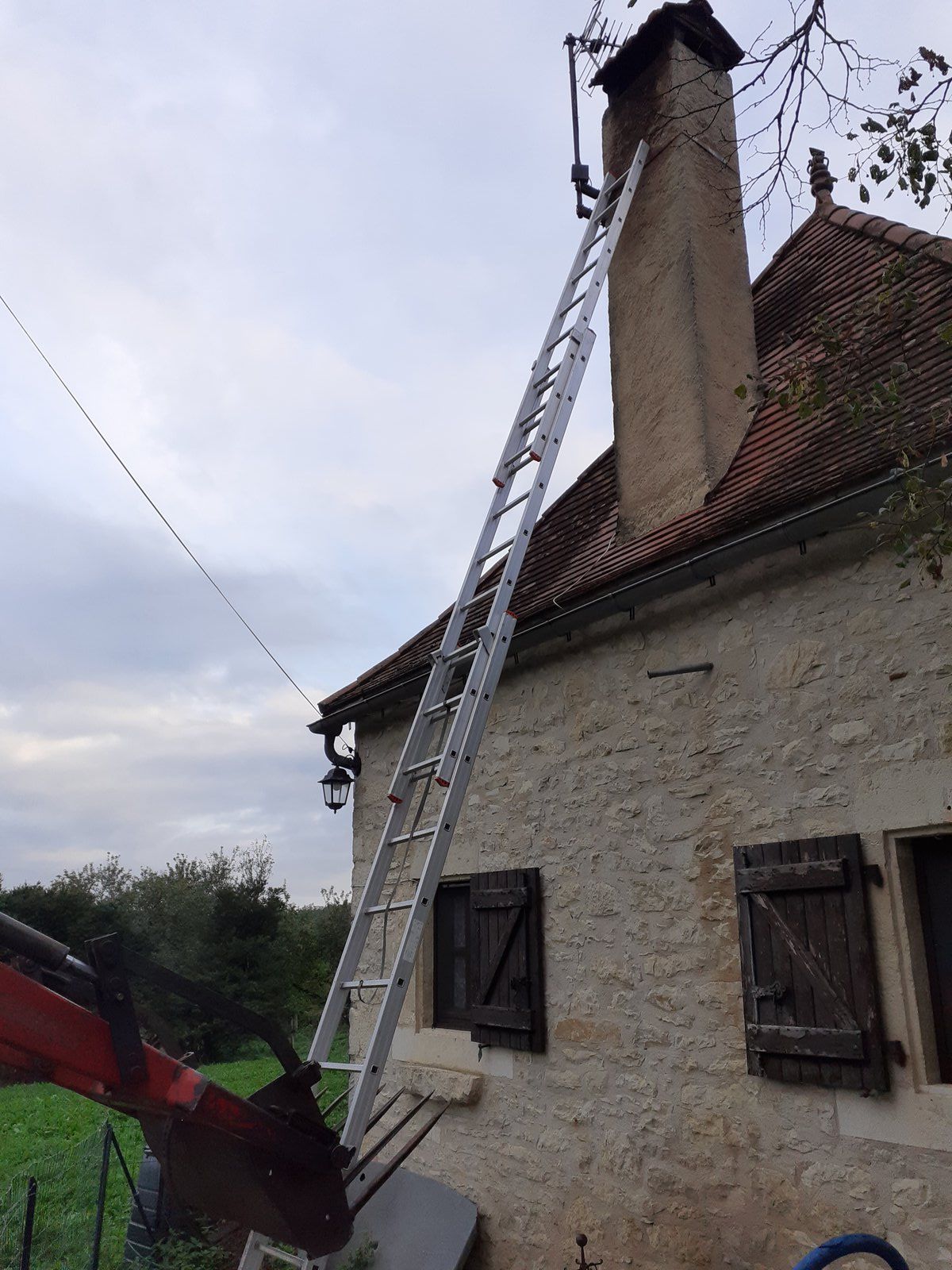 Échelle appuyée contre la cheminée d'une maison en pierre, avec un ascenseur ou une machinerie rouge à gauche.