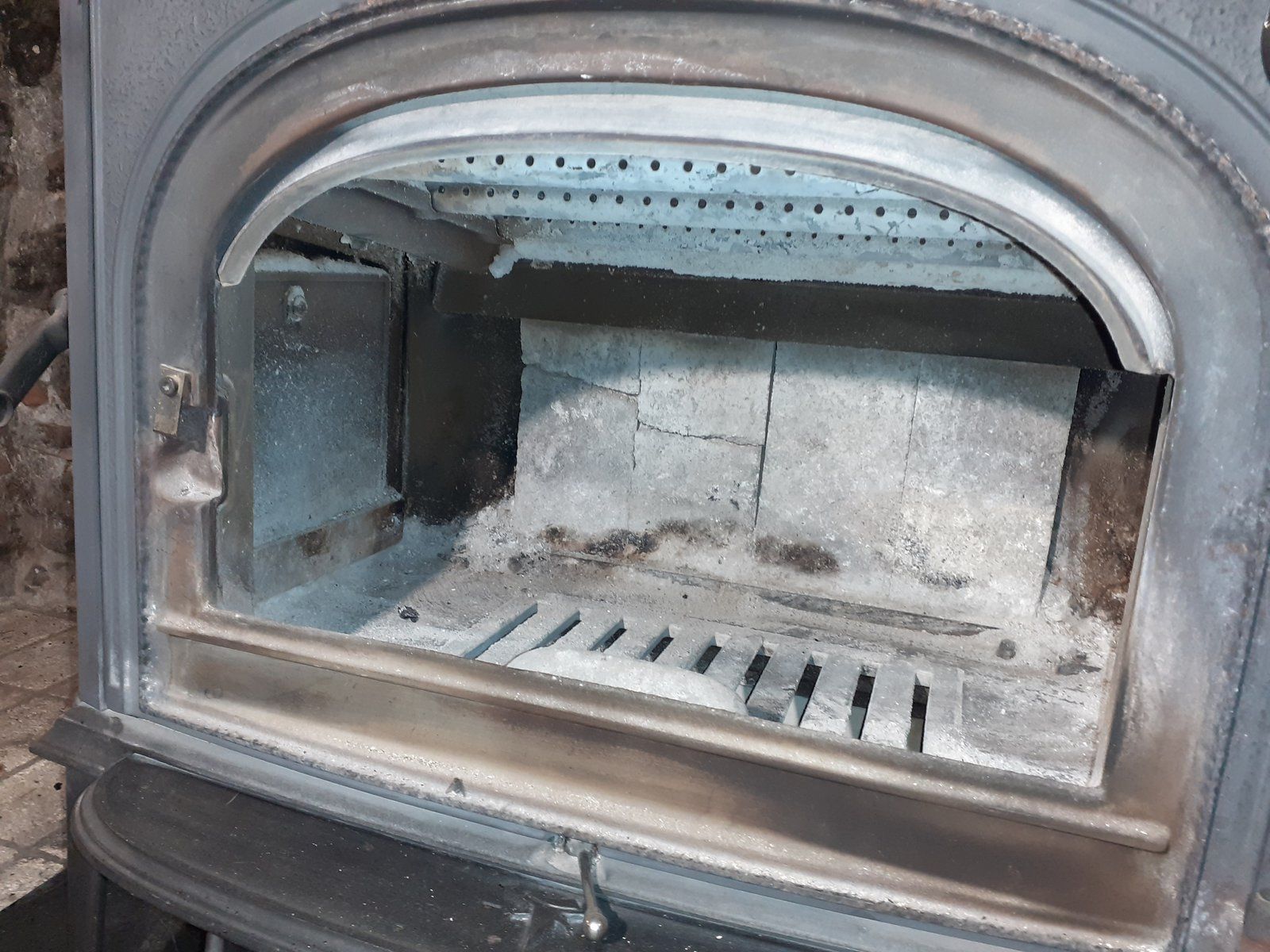 Intérieur d'un petit poêle à bois ou d'une cheminée, avec des résidus de cendres et une porte noircie par la suie, ouverte.
