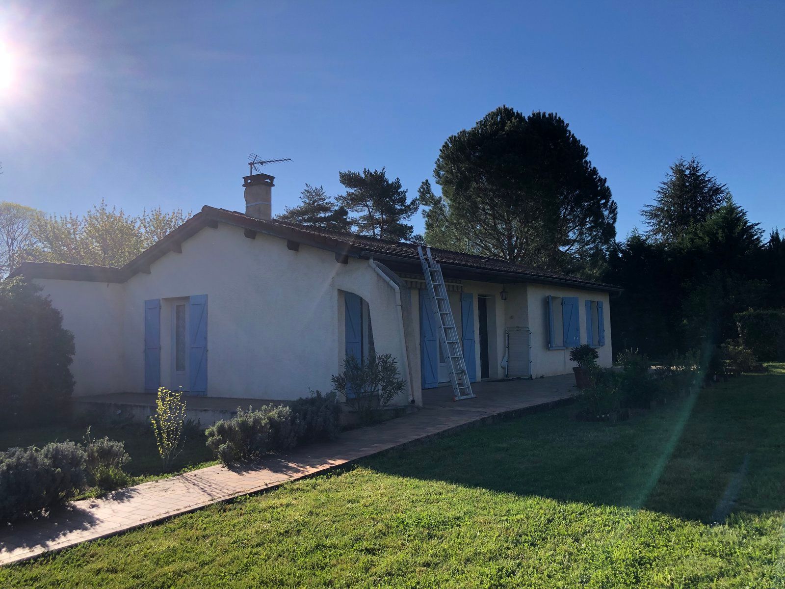 Maison de plain-pied avec volets bleus, porche et pelouse verte baignée de soleil.