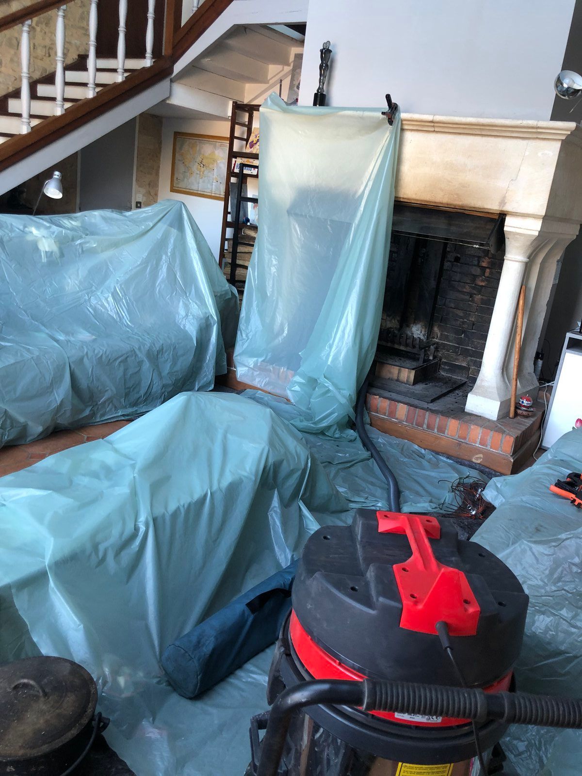 Salon recouvert de bâches en plastique, avec une cheminée et un aspirateur rouge et noir au premier plan.
