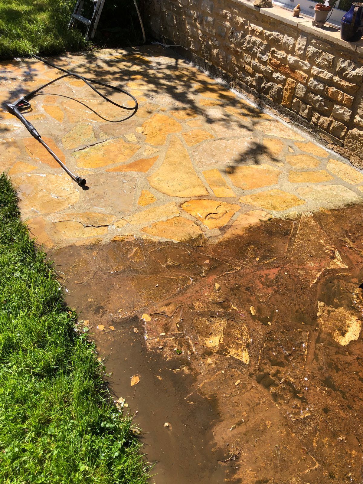 Terrasse arrière avec lance de nettoyeur haute pression et eaux boueuses ruisselant sur une dalle de béton près d'un mur en pierre