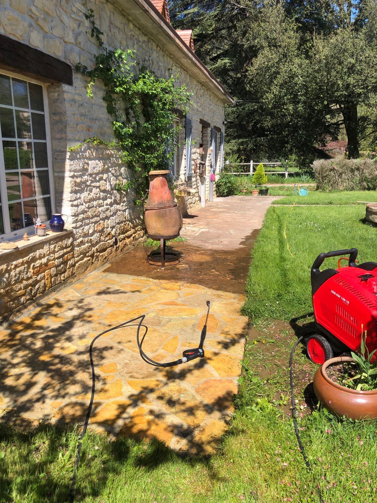 Terrasse en pierre jouxtant une maison avec un mur recouvert de vigne, une pelouse et un barbecue rouge au soleil.