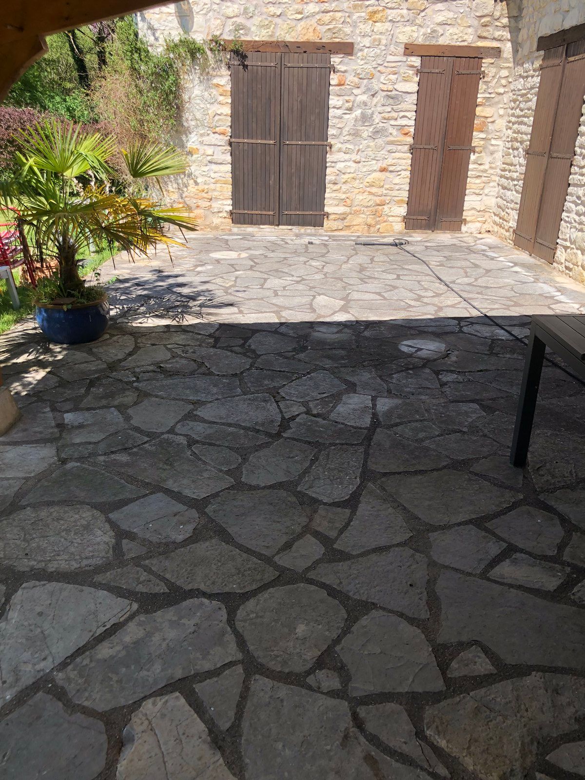 Cour-terrasse en pierre avec portes en bois, plantes en pot et mur en pierre baigné de soleil.