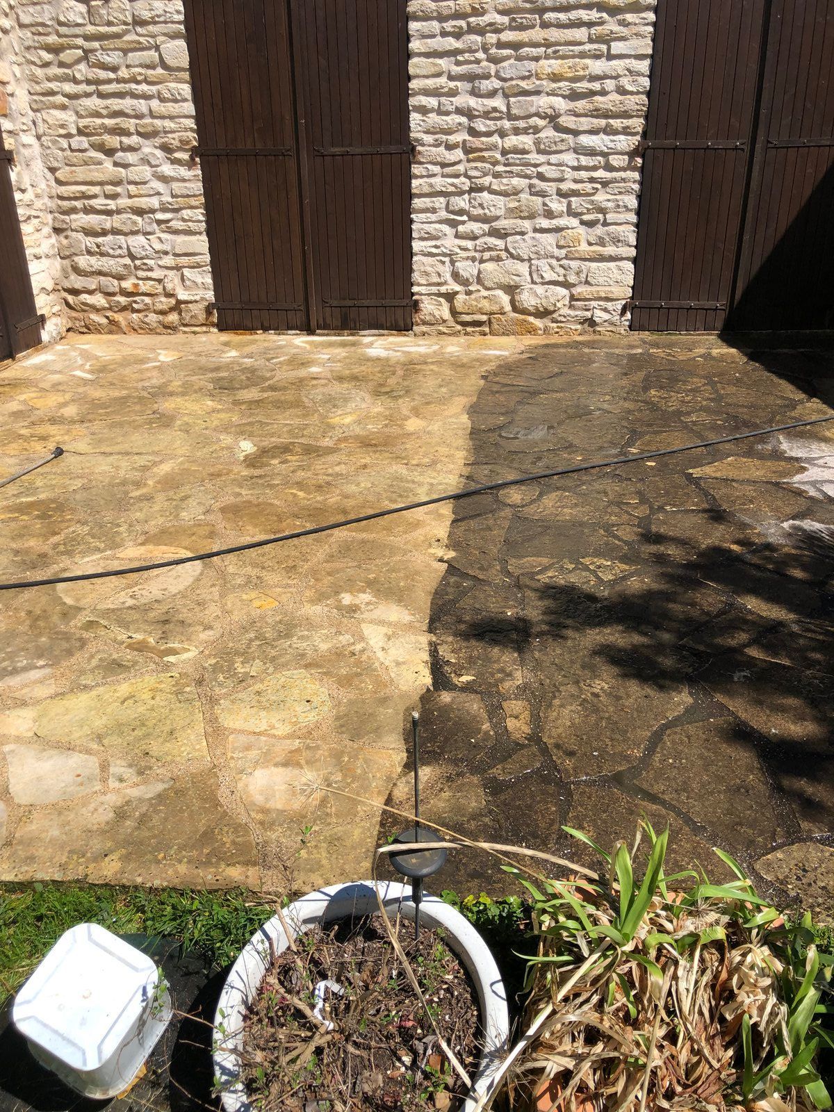 Terrasse en pierre adossée à un mur de briques avec des portes en bois foncé, un pot de fleurs et un petit récipient blanc au premier plan.