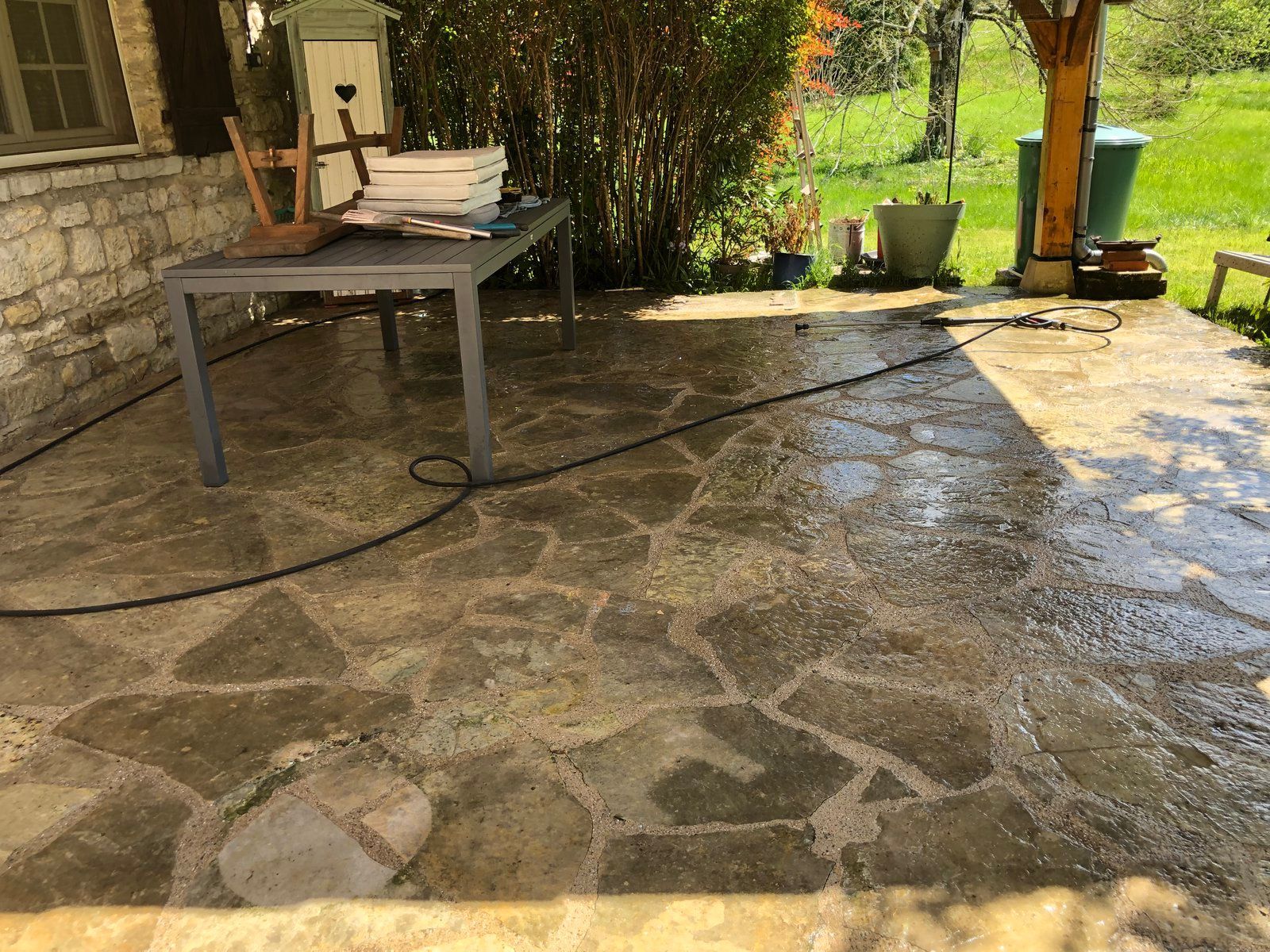 Terrasse extérieure avec sol en pierre, table en métal et tuyau d'arrosage près d'un muret en pierre et d'arbres