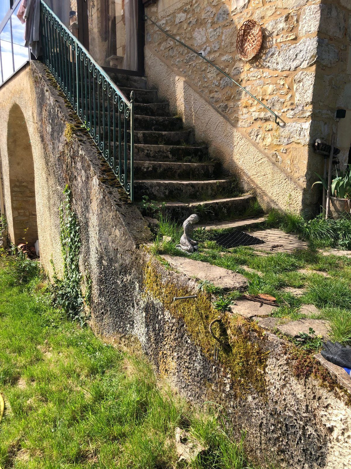 Un escalier extérieur en pierre longe un vieux mur, avec une rampe noire et des touffes d'herbe verte.