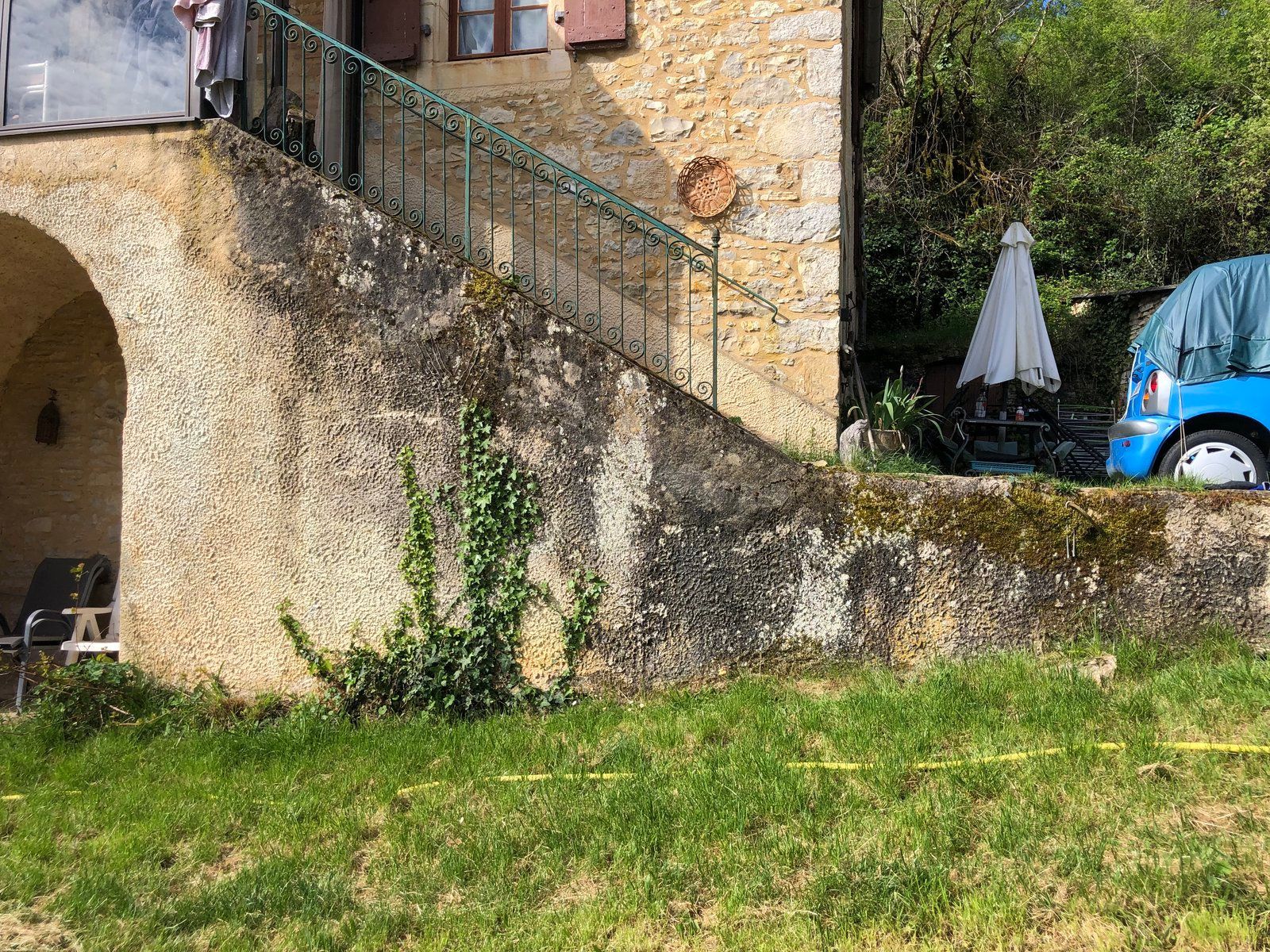 Mur de terrasse en pierre avec escalier, premier plan herbeux et voiture bleue garée près d'un coin de jardin ombragé.