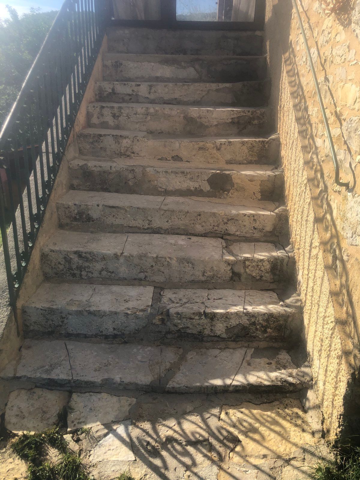 Escalier extérieur en béton à forte pente, adossé à un mur jaune et une rampe noire, baigné de soleil.