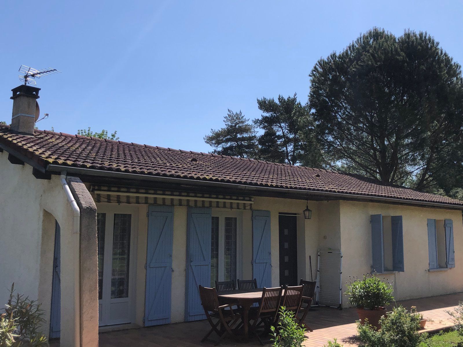 Maison rustique baignée de soleil, avec volets bleus, toit de tuiles et table de patio sous un grand arbre.