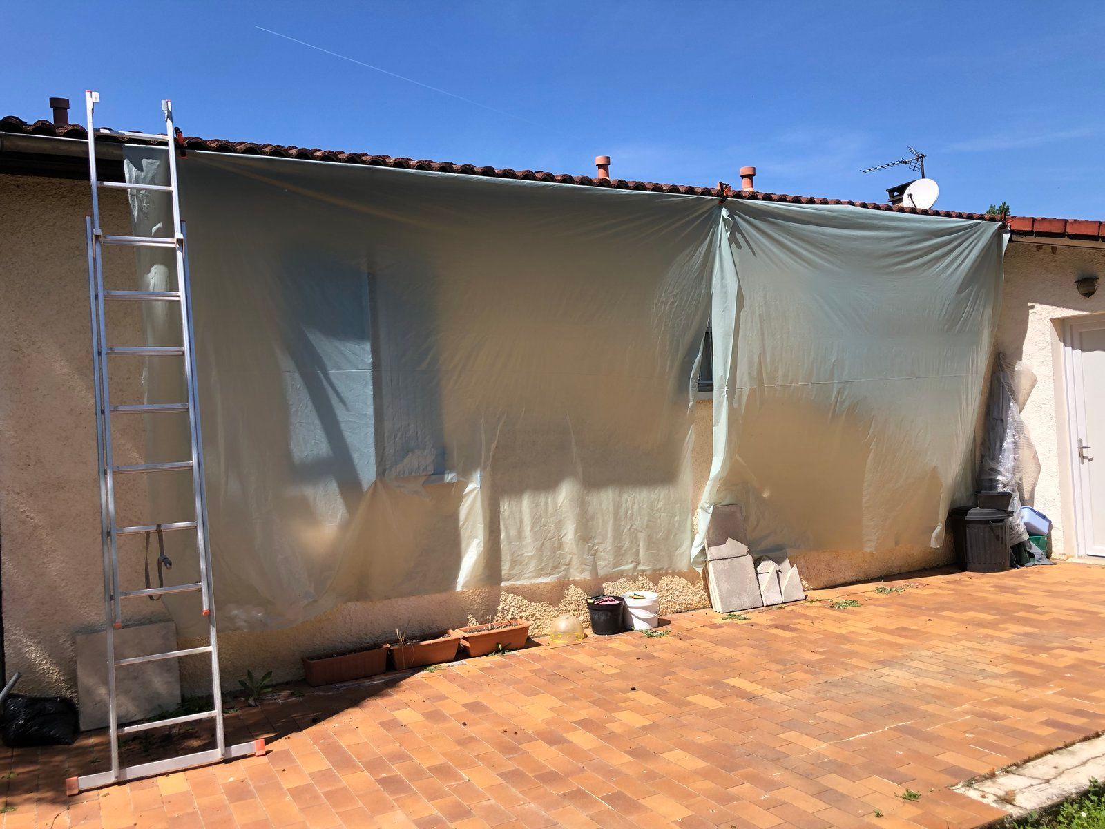 Terrasse extérieure avec une échelle contre un mur de maison recouvert de bâches en plastique suspendues sous un ciel bleu