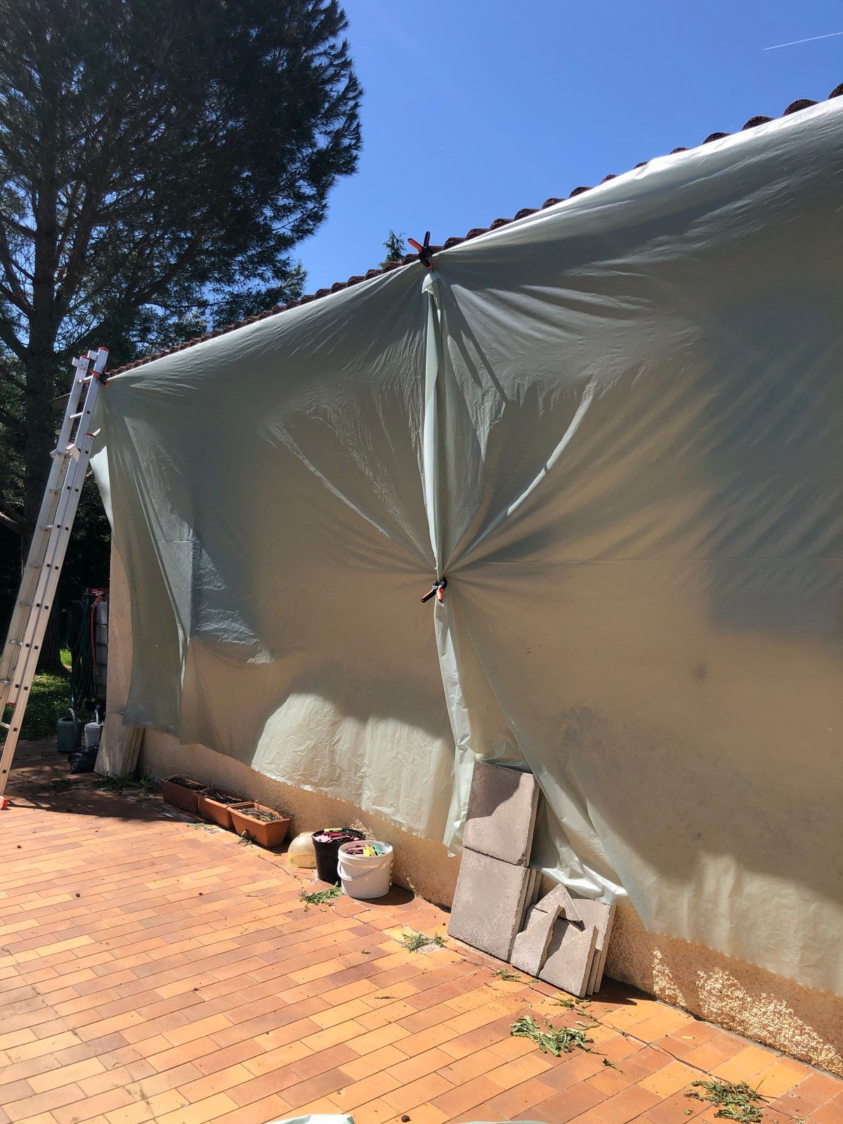 Structure recouverte d'une bâche blanche à côté d'une terrasse en briques sous un ciel bleu, avec des arbres et des poteaux de soutien à proximité.