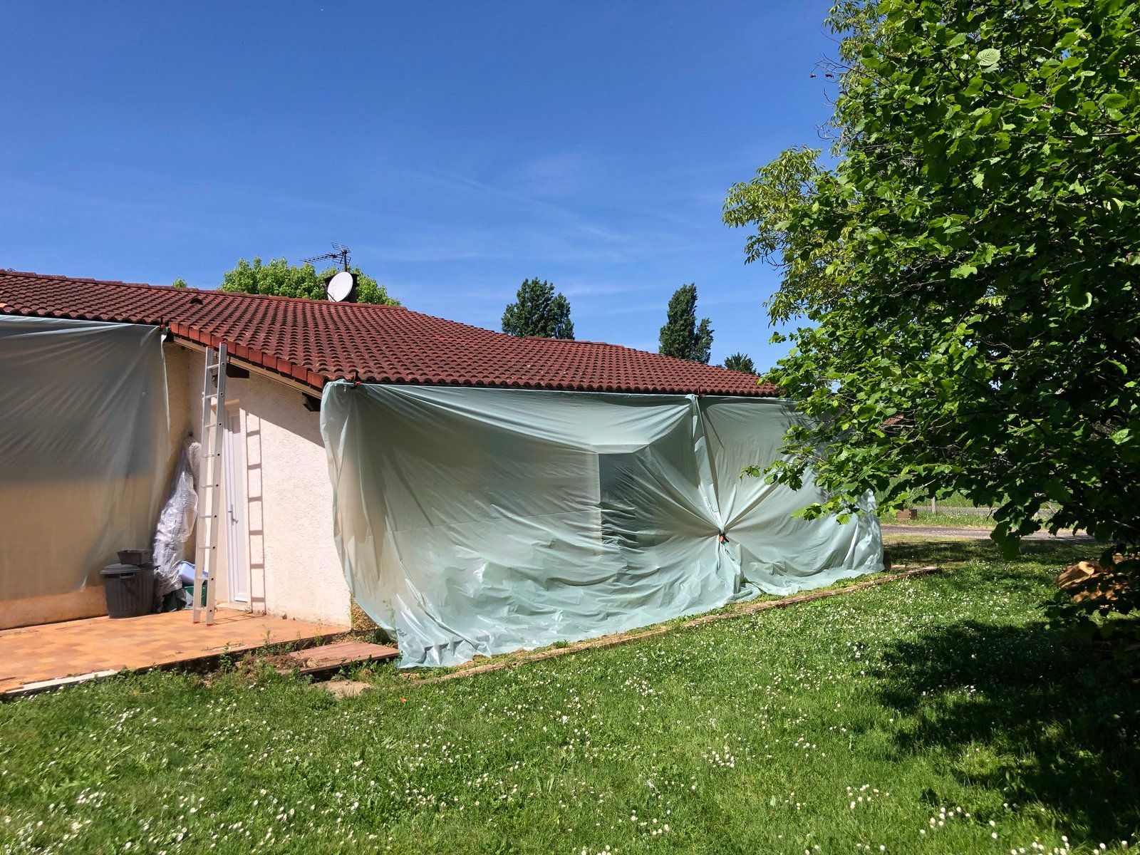 Maison au toit de tuiles rouges recouverte d'une bâche en plastique transparent, sous un ciel bleu et à côté d'une pelouse verte