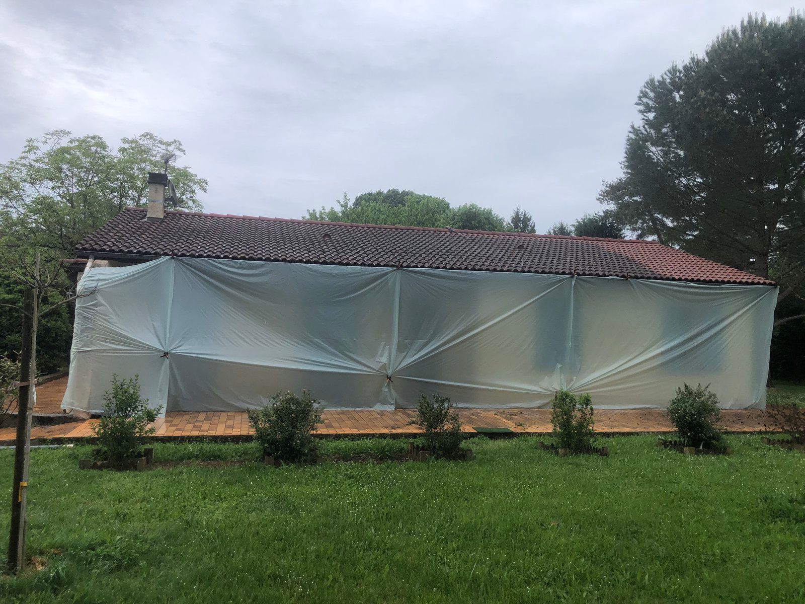 Petite maison au toit de tuiles rouges recouvert d'une bâche blanche, derrière une cour herbeuse et des arbustes sous un ciel nuageux