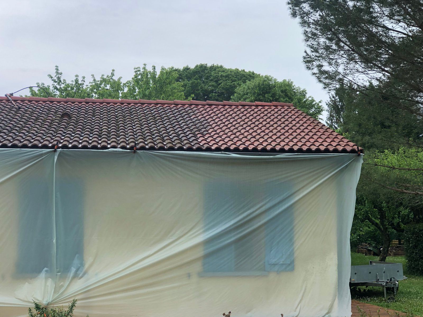 Maison au toit de tuiles rouges recouverte d'une bâche de protection en plastique à l'extérieur, avec des arbres derrière.