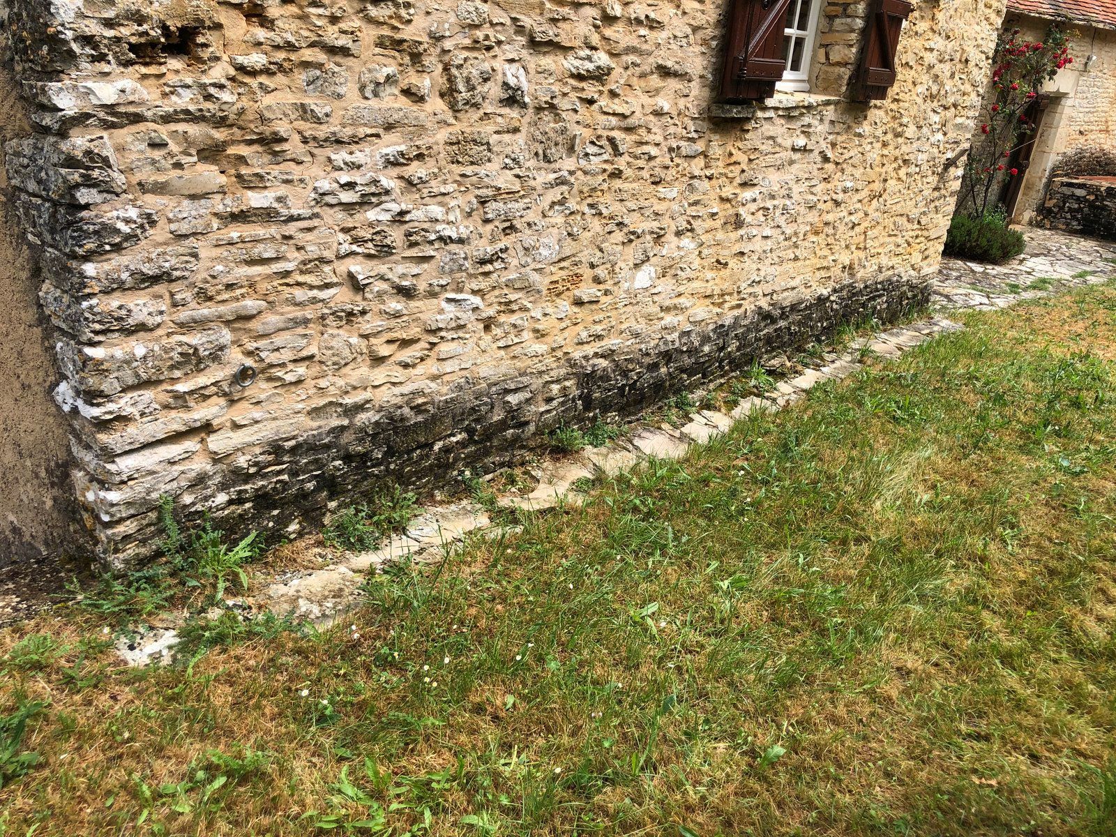 Mur en pierre bordant une bande herbeuse, avec une petite fenêtre et une maçonnerie brute et patinée par le temps.