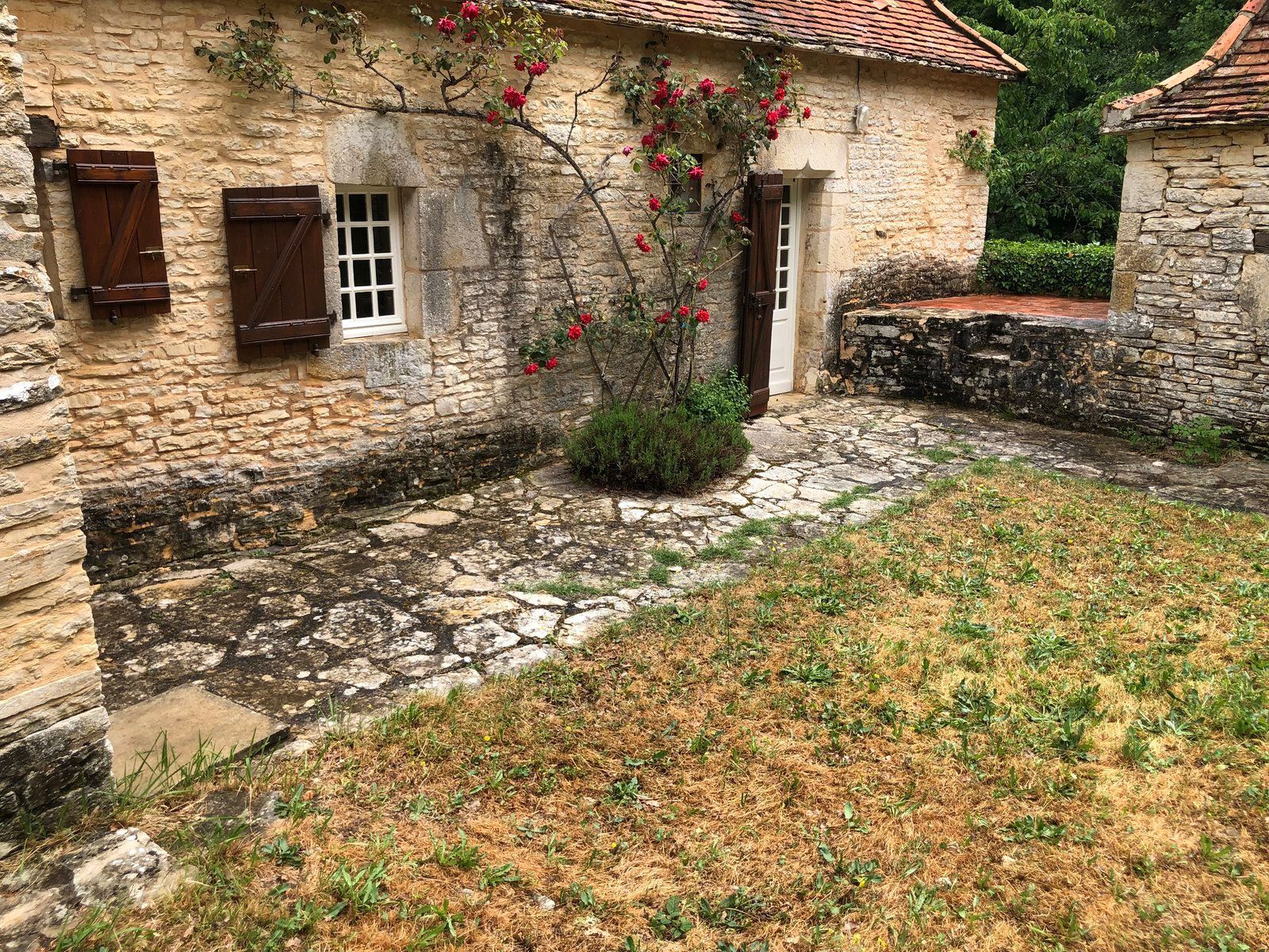 Cour intérieure d'une maison en pierre avec fenêtres blanches, toit de tuiles rouges et mur recouvert de vigne donnant sur une pelouse