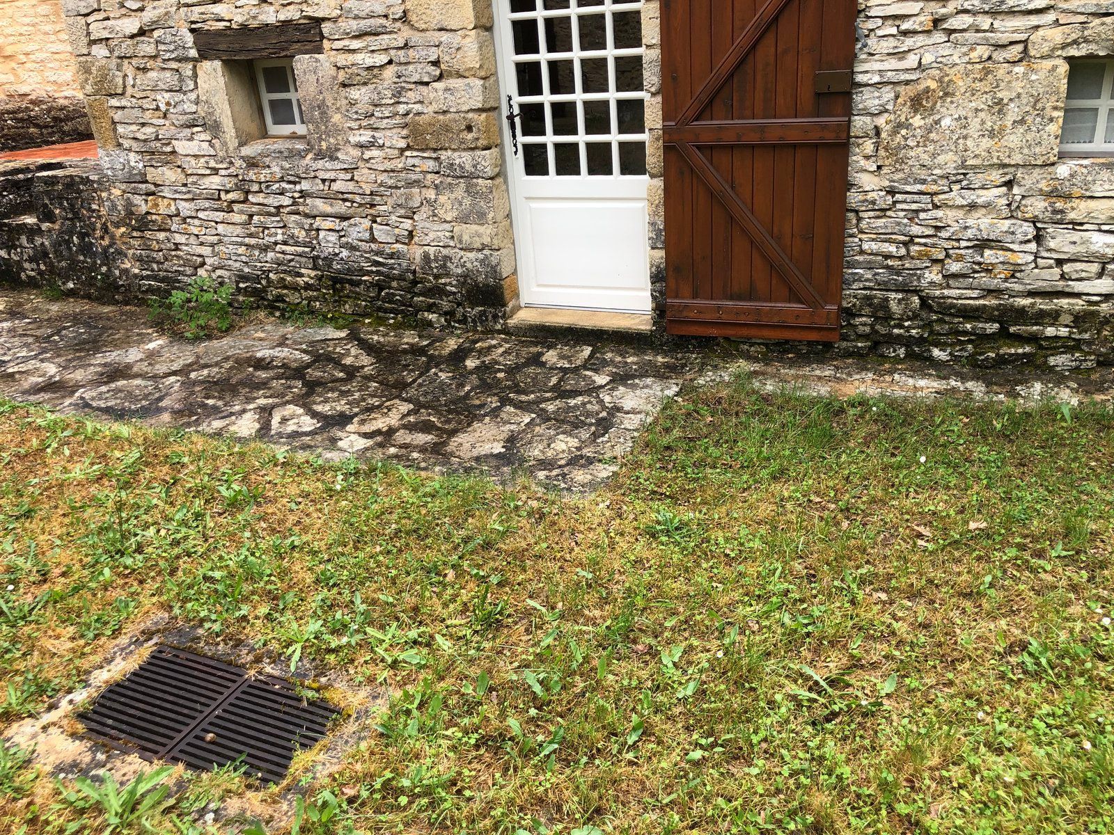 Mur en pierre d'une maison avec porte blanche, portail en bois et cour gazonnée avec grille d'égout.
