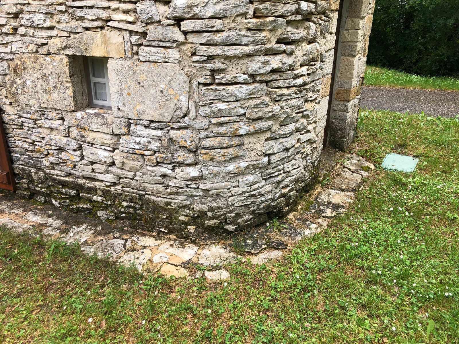 Coin d'un bâtiment en pierre avec une petite fenêtre et un terrain herbeux tout autour.