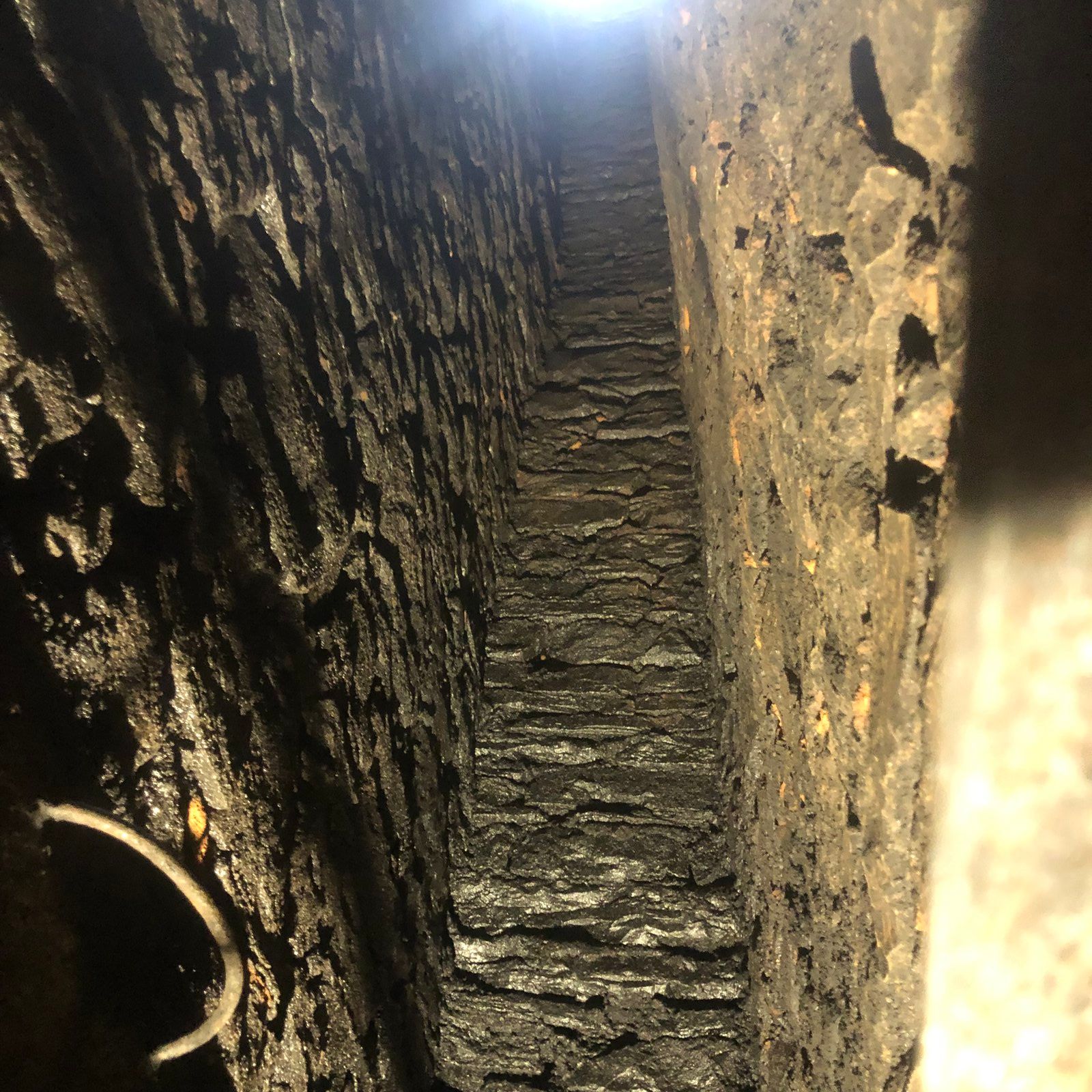 Tunnel étroit en pierre aux parois humides, éclairé par le haut, avec un tuyau longeant le côté gauche.