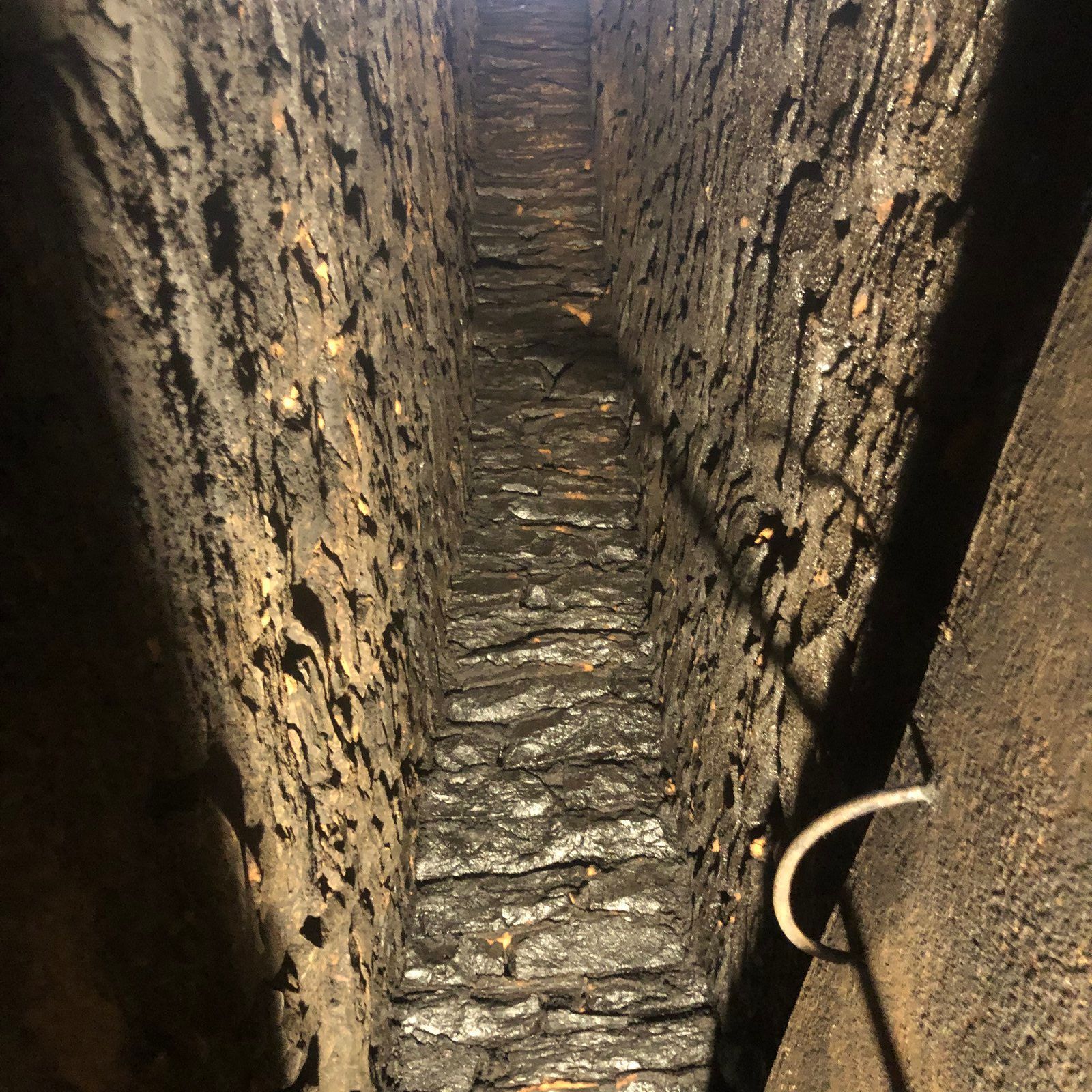 Étroite crevasse rocheuse au sol de pierre rugueux et humide, avec un crochet métallique sur le bord droit.