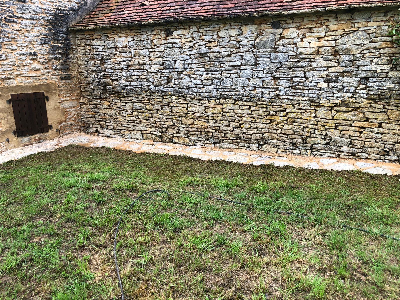 Mur en pierre avec une petite fenêtre donnant sur une cour gazonnée et un toit de tuiles.
