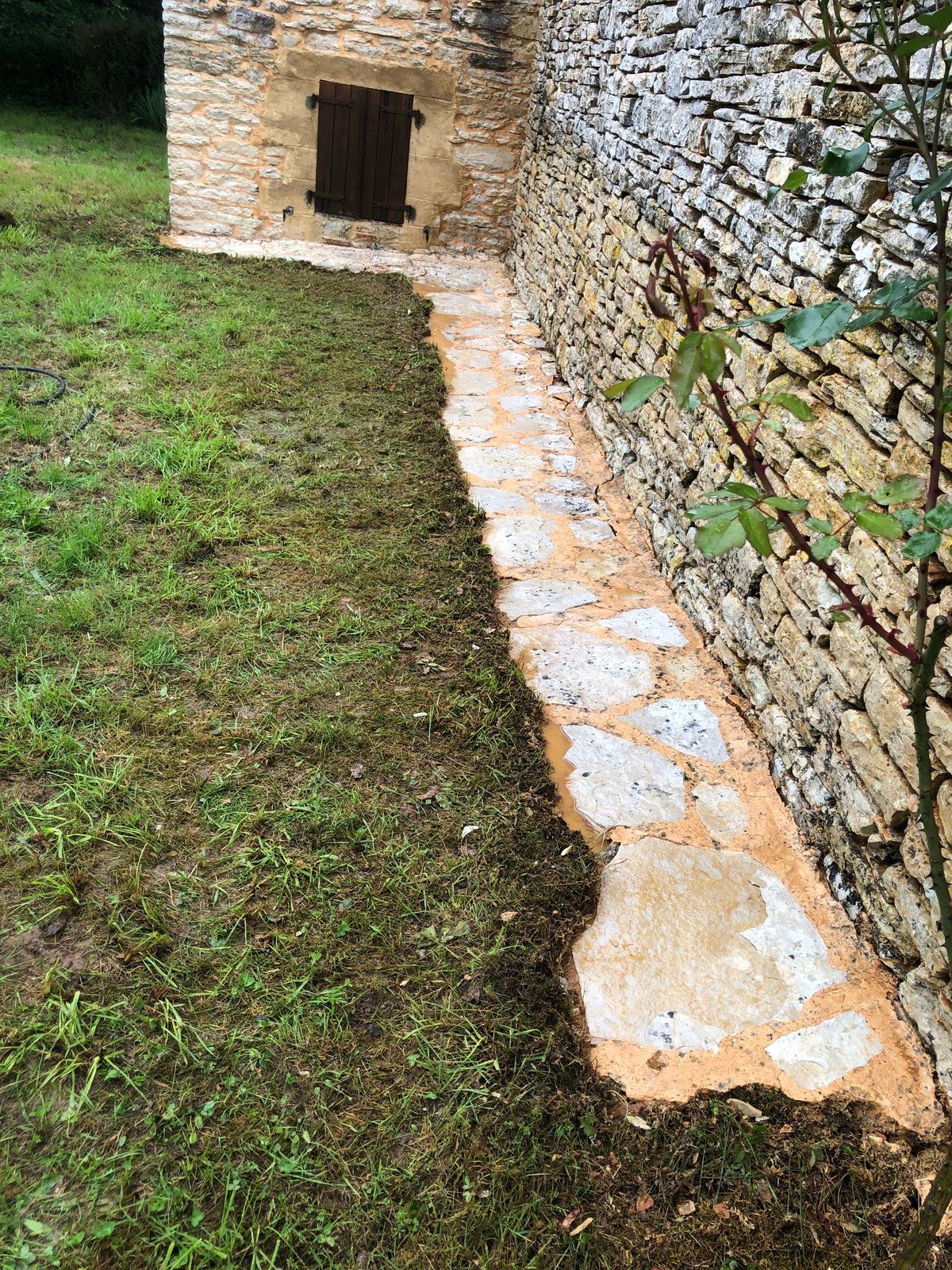 Canal de drainage en pierre longeant une pente herbeuse et un mur de pierre, menant à une petite ouverture dans le mur.