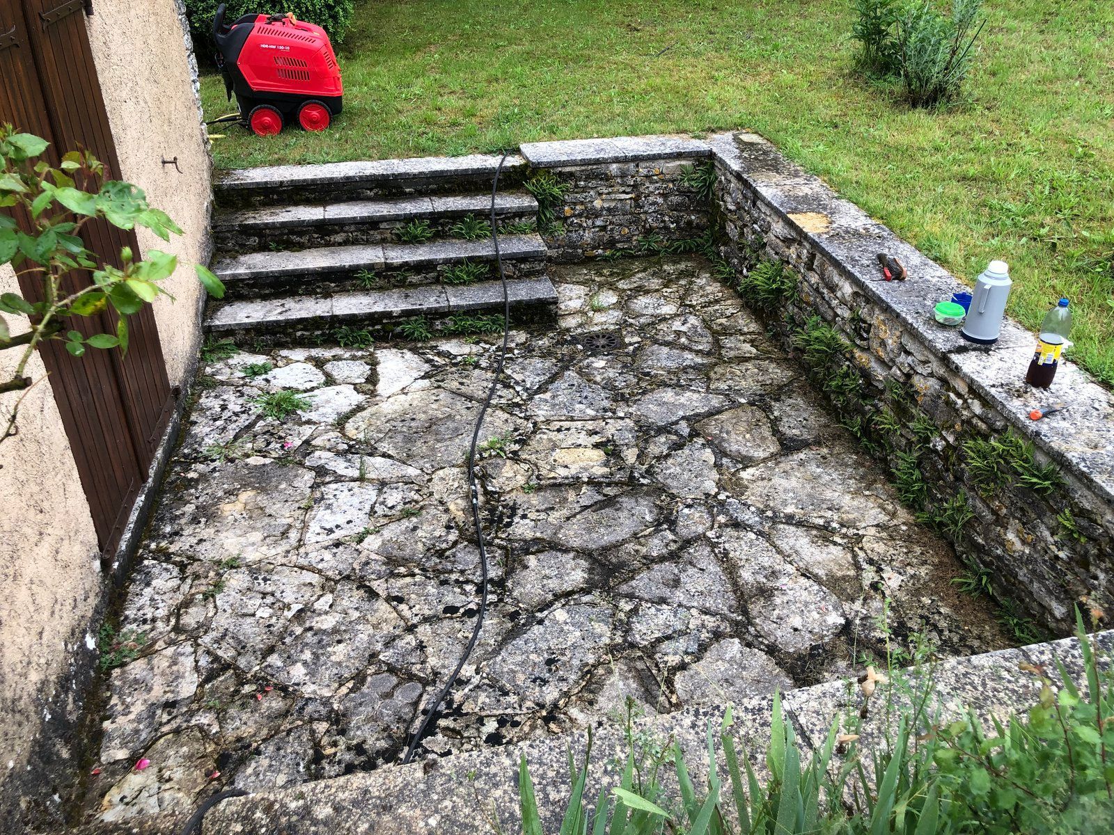 Terrasse en béton fissuré avec marches, bordée de gazon et d'un muret en pierre, avec une tondeuse à gazon rouge dans la cour.