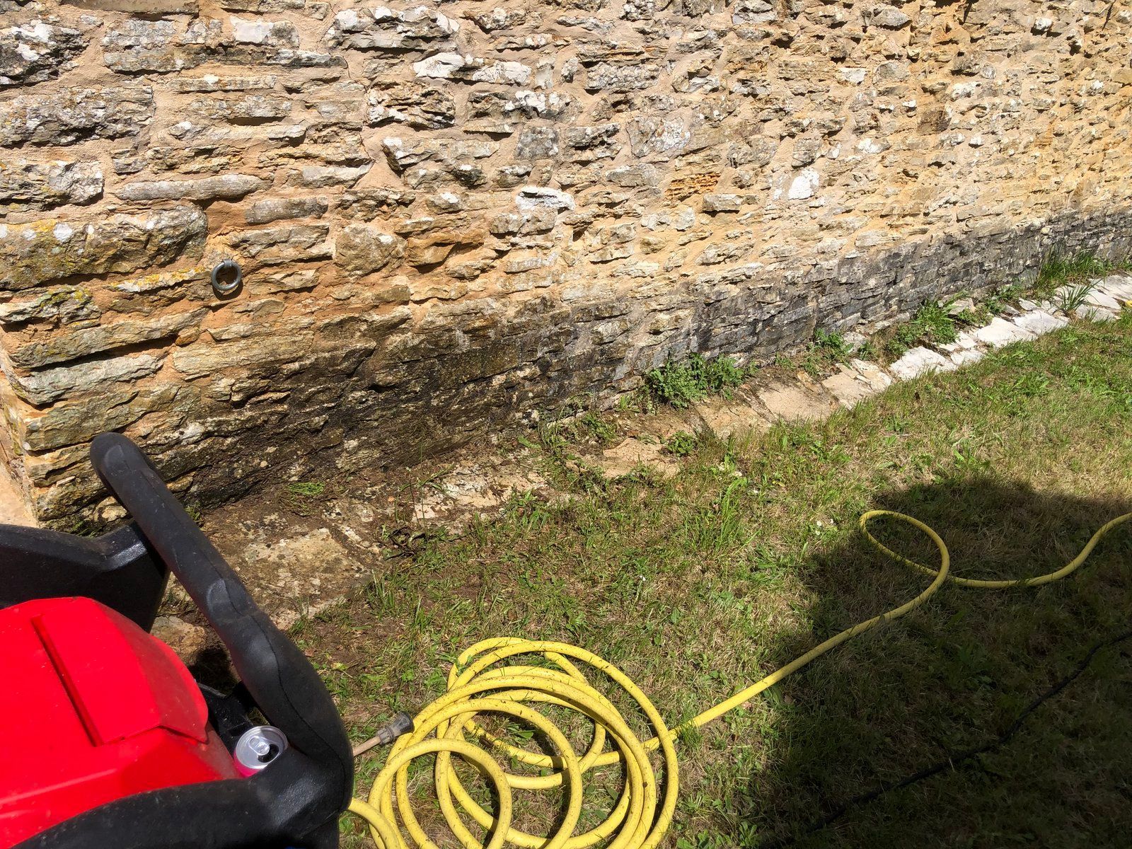 Outil de jardinage rouge avec tuyau jaune enroulé sur l'herbe près d'un mur de pierre