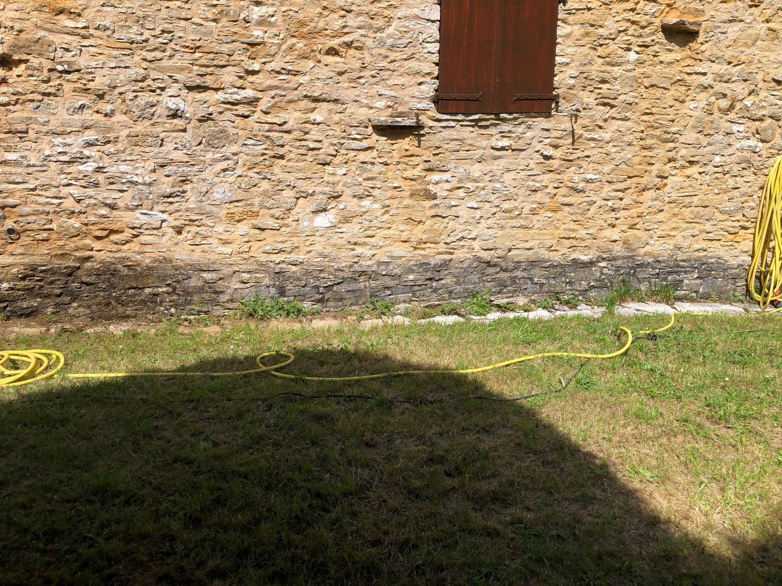 Un mur de pierre et une porte rouge surplombent une cour herbeuse où se dessine une grande ombre au premier plan.