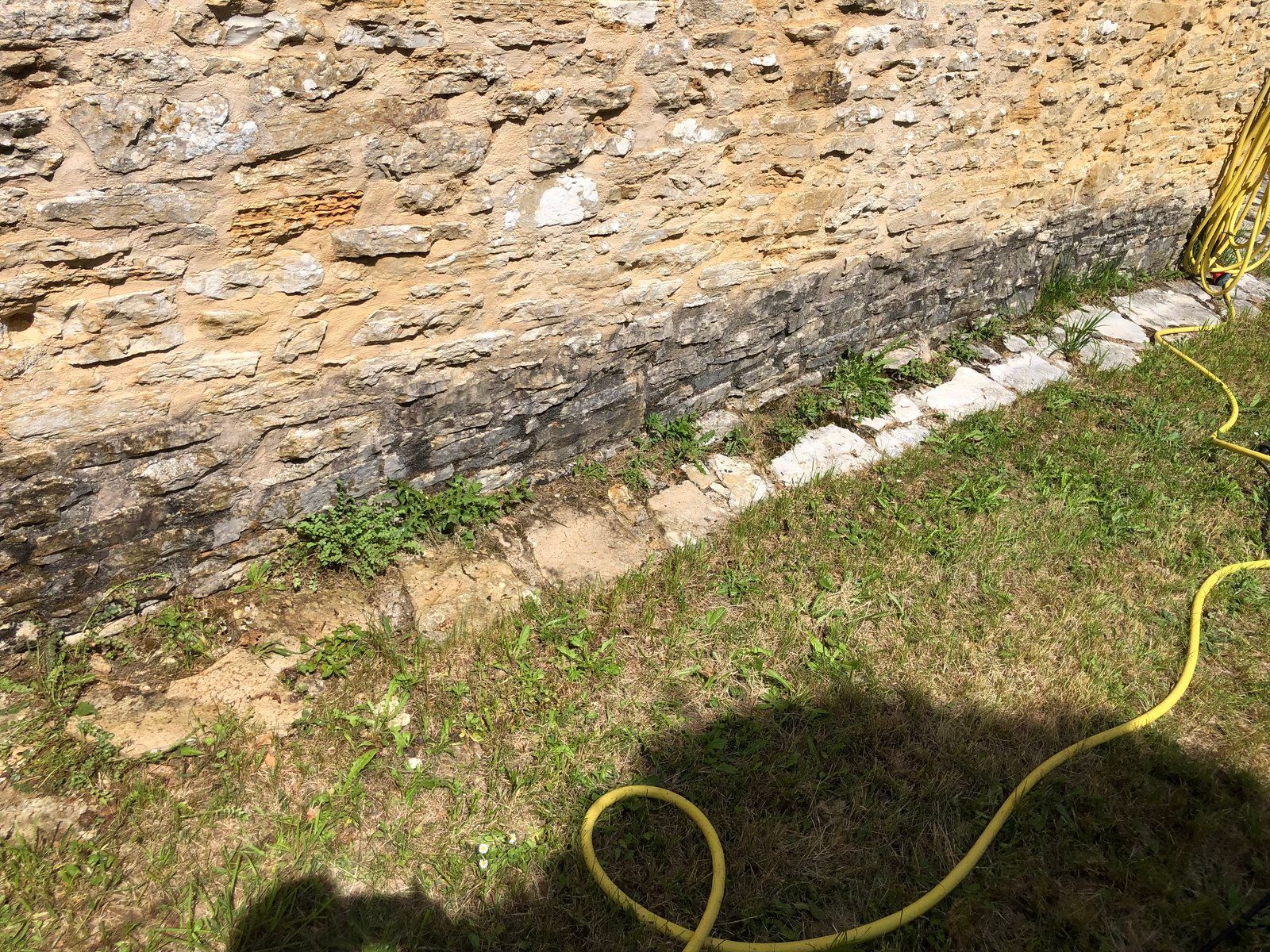 Tuyau jaune sur l'herbe, à côté d'un muret en pierre brute et d'un rebord étroit en béton.