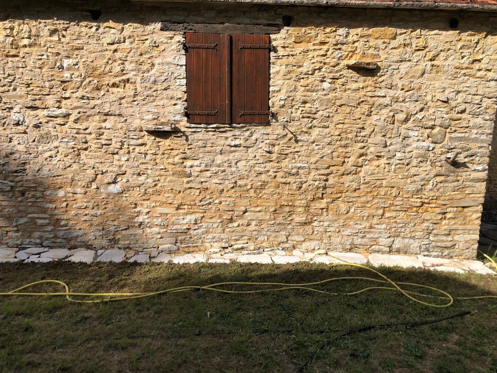 Mur en pierre avec une petite fenêtre à volets bruns donnant sur une cour gazonnée.