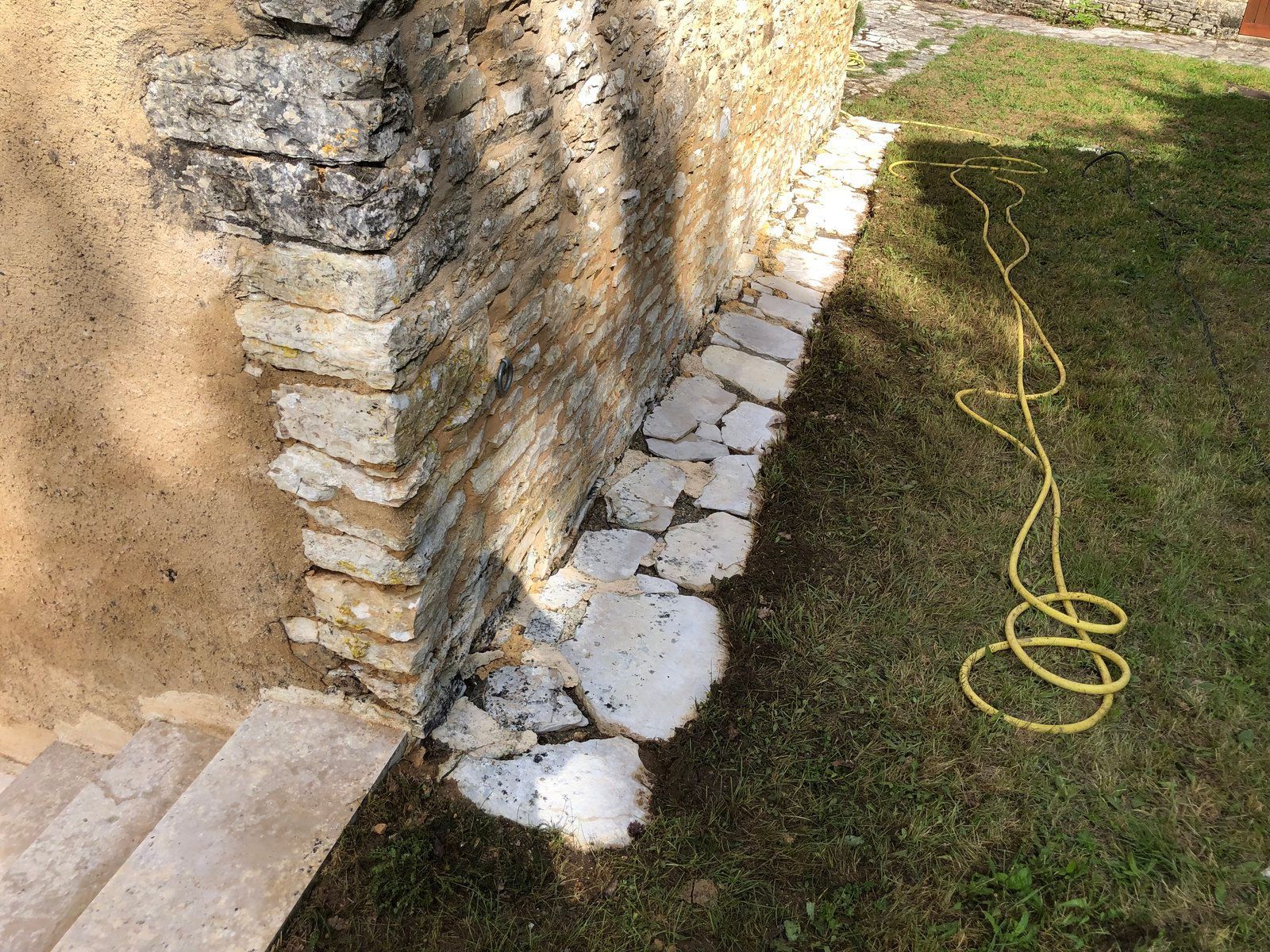 Coin d'une maison en pierre avec des briques empilées, des marches et un tuyau d'arrosage jaune sur l'herbe.