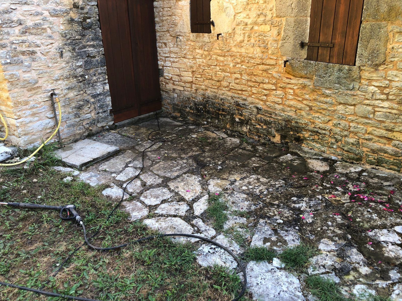 Cour en pierre jouxtant un mur de briques patinées et une porte en bois, avec des touffes de mousse et d'herbe.