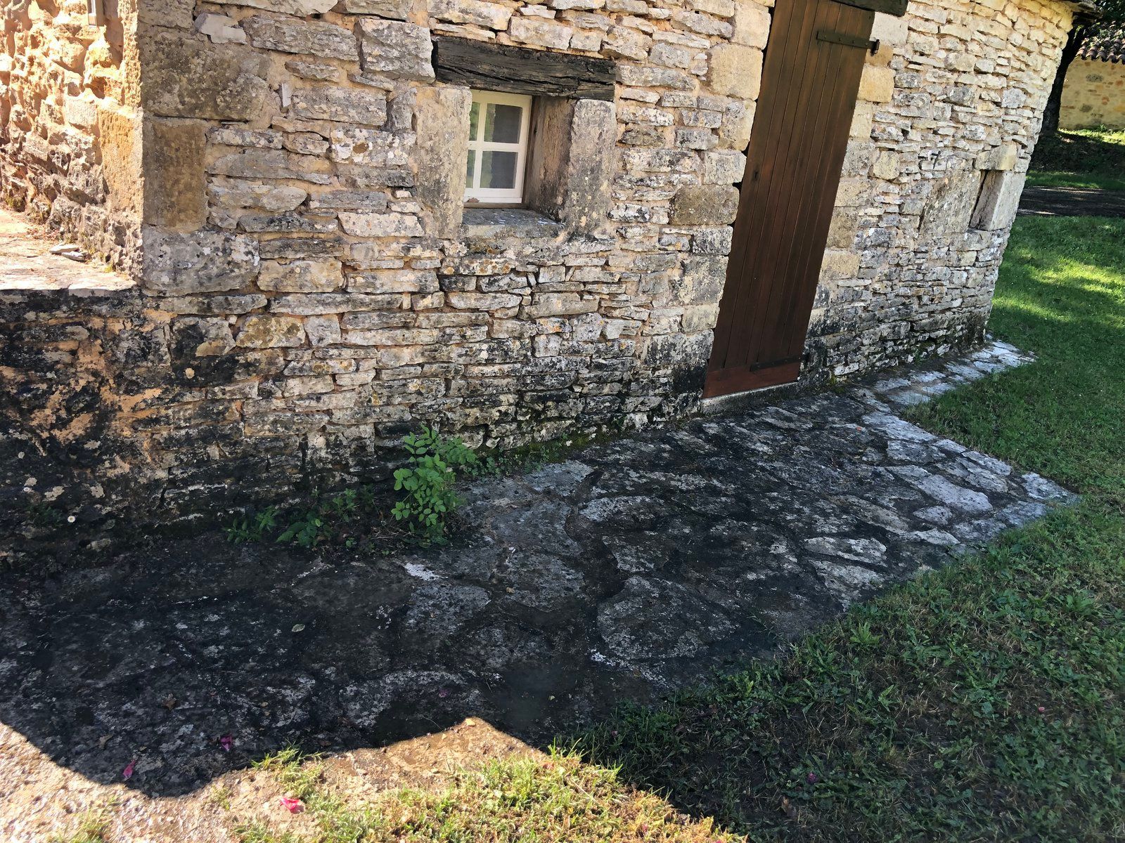 Mur en pierre d'une maisonnette avec une petite fenêtre, une porte en bois et un chemin moussu bordant l'herbe