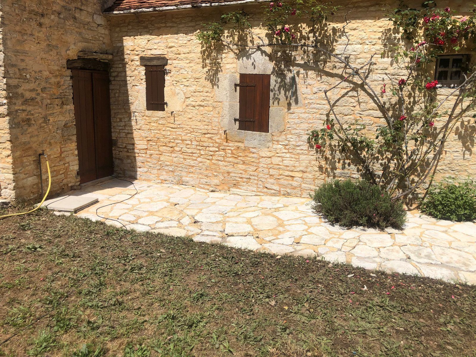 Cour en pierre avec une porte en bois, des volets et une vigne fleurie sur un mur rustique baigné de soleil