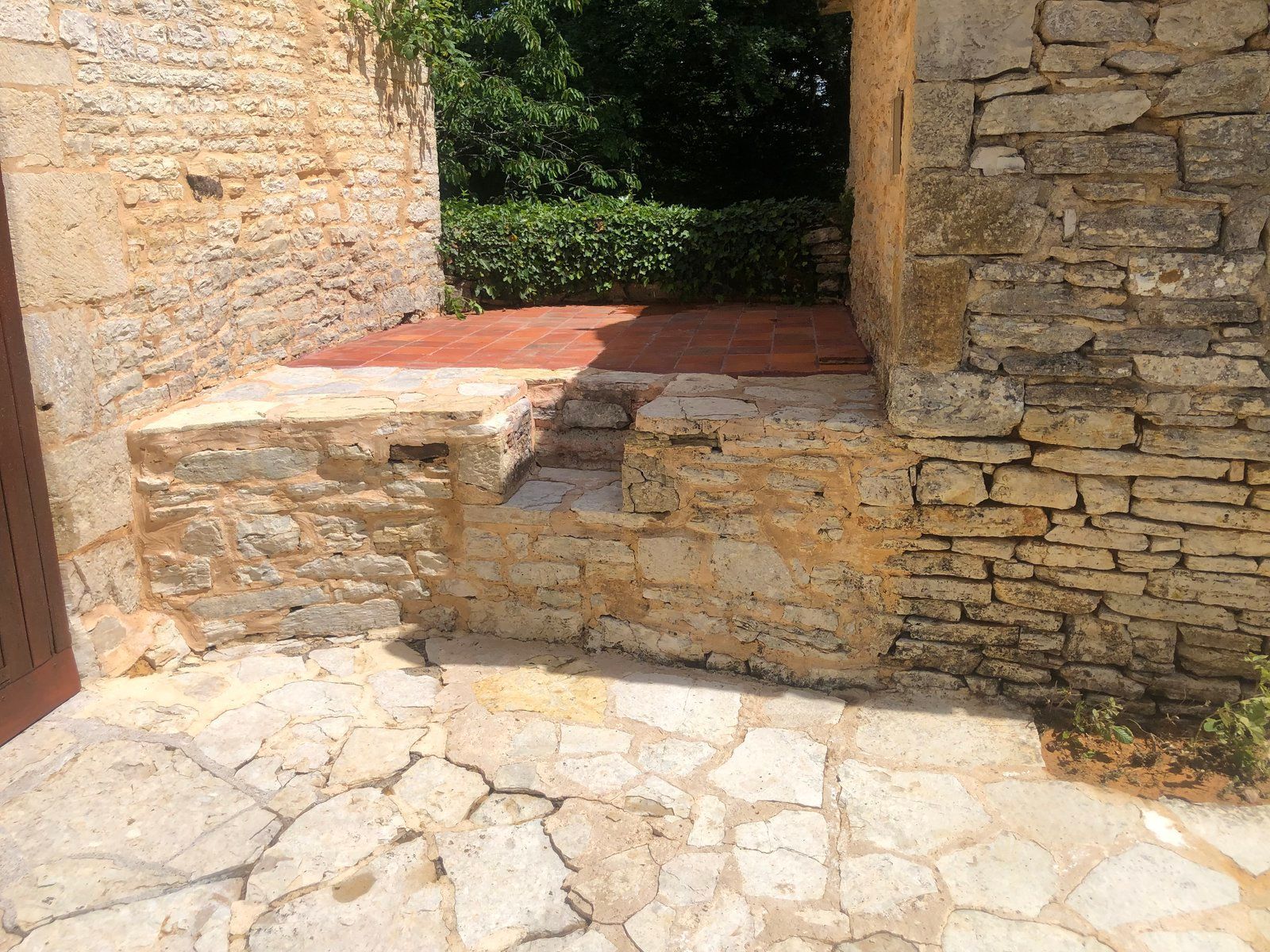 Des marches en pierre et une petite terrasse ensoleillée entre des murs de pierre patinés par le temps.