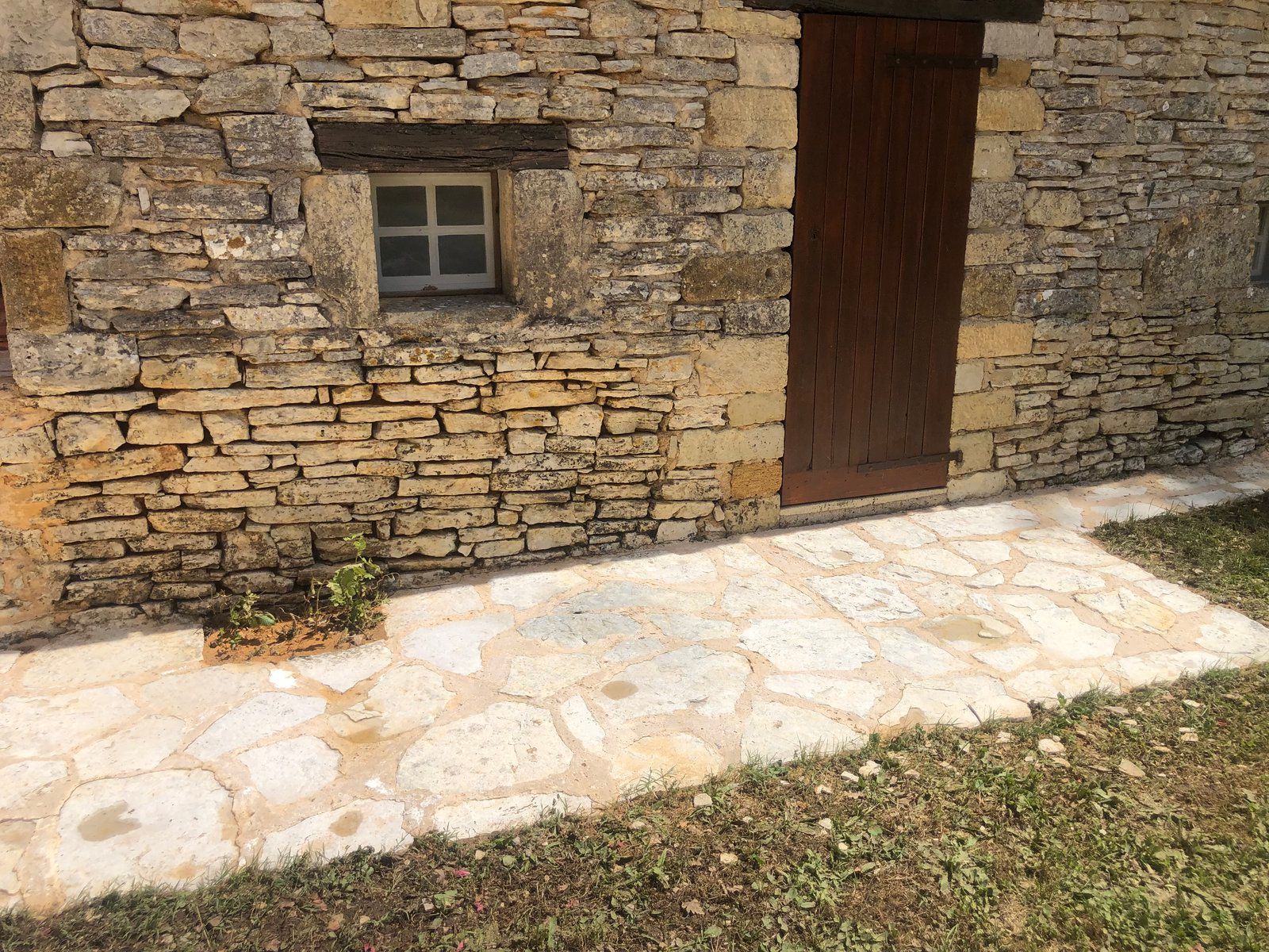 Mur en pierre avec une petite fenêtre, porte en bois et chemin pavé bordant l'herbe.