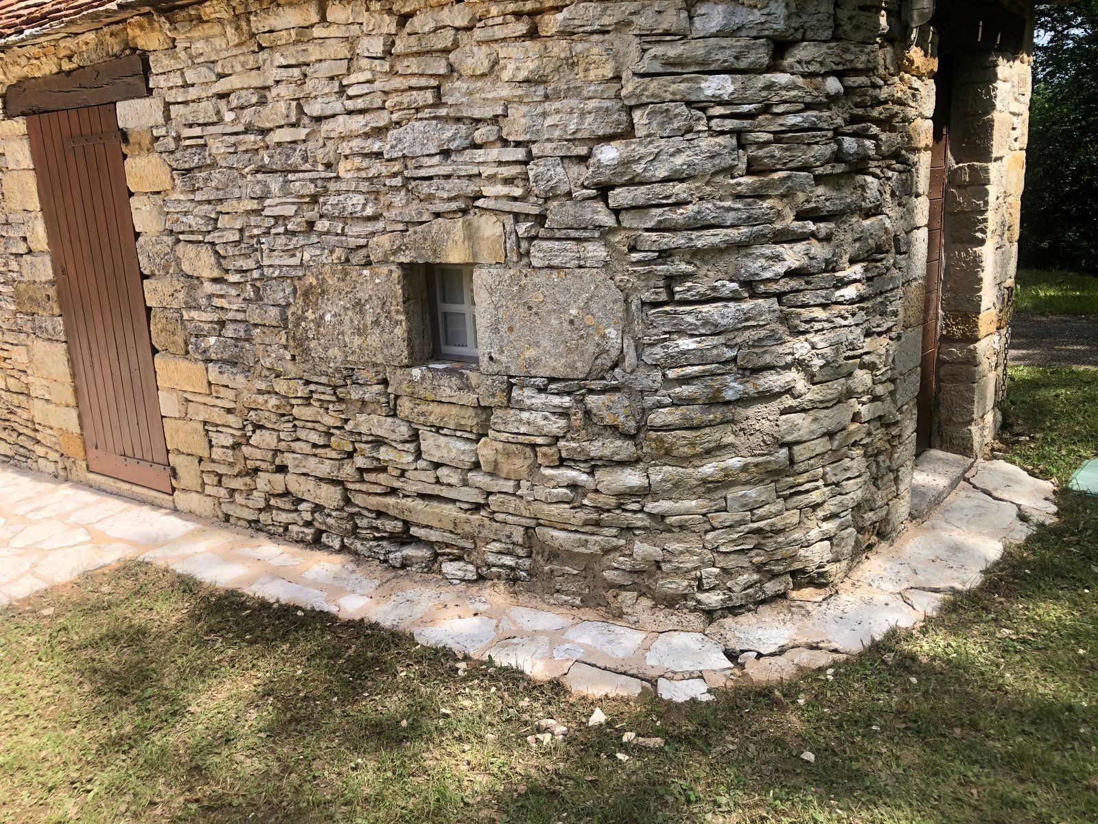 Bâtiment en pierre d'angle avec une petite fenêtre, une porte en bois et un chemin pavé bordant l'herbe.