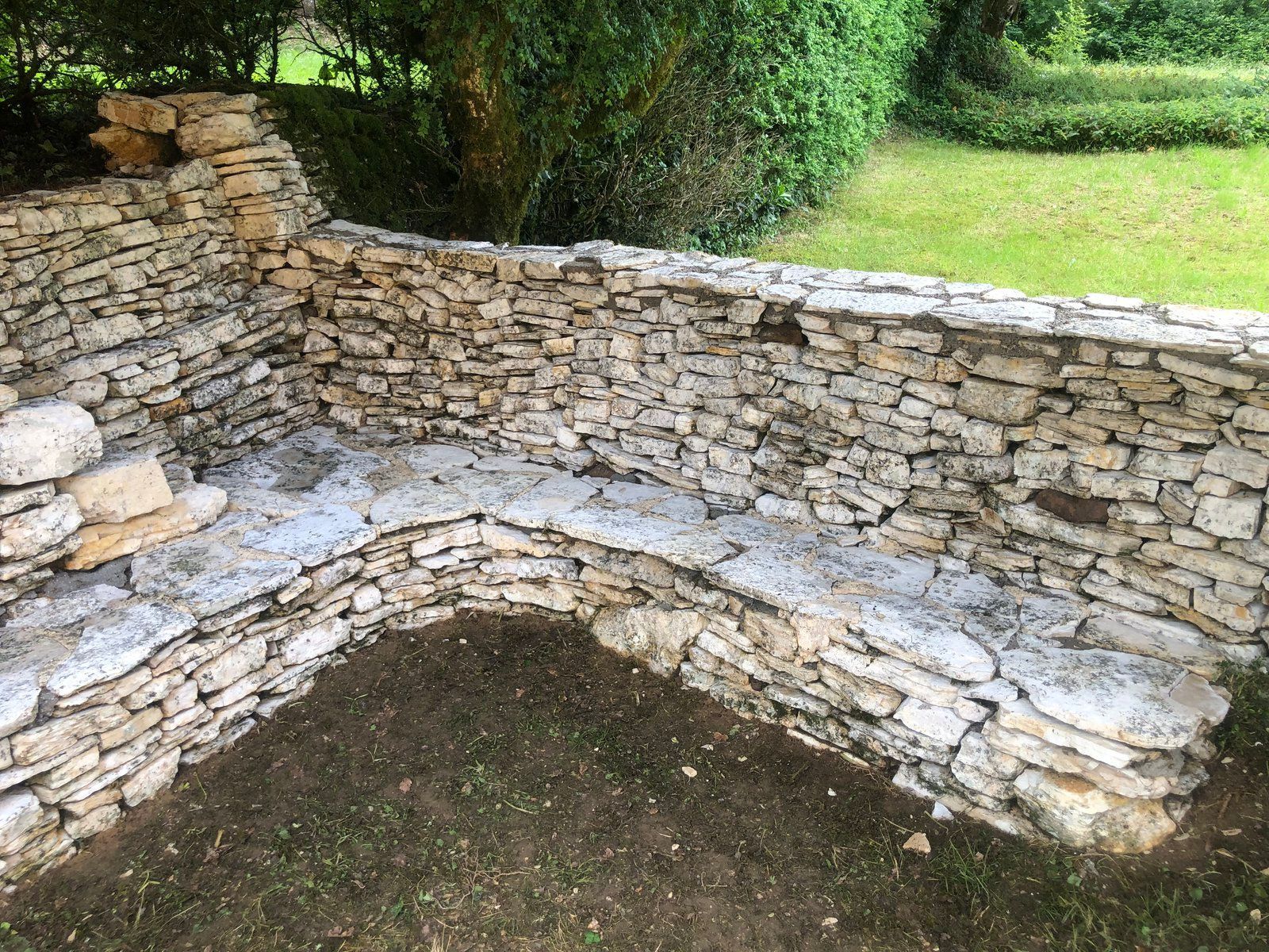 Mur de jardin en pierre avec un banc incurvé et un sol en terre battue dans un espace extérieur arboré