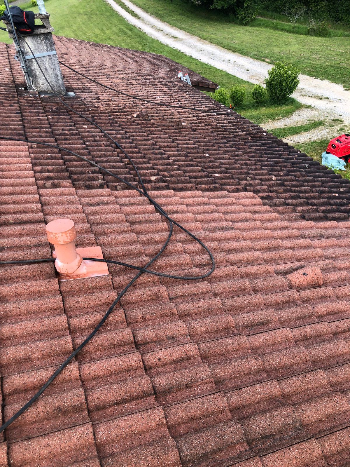 Toiture en cours de réparation avec des câbles noirs sur des bardeaux rougeâtres ; cheminée visible près d'une cour gazonnée.