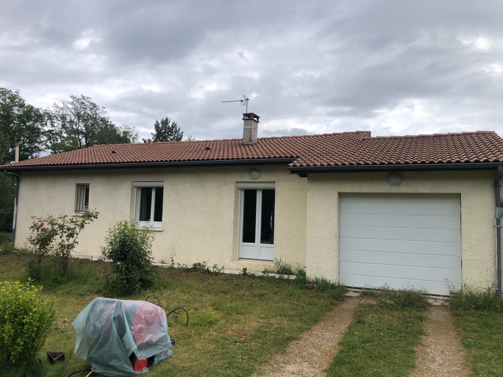 Maison de plain-pied avec toit de tuiles rouges, garage et jardin gazonné sous un ciel nuageux