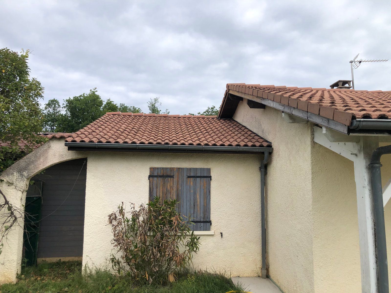 Petite maison couleur crème, au toit de tuiles rouges et à la fenêtre fermée par des volets, sous un ciel nuageux.