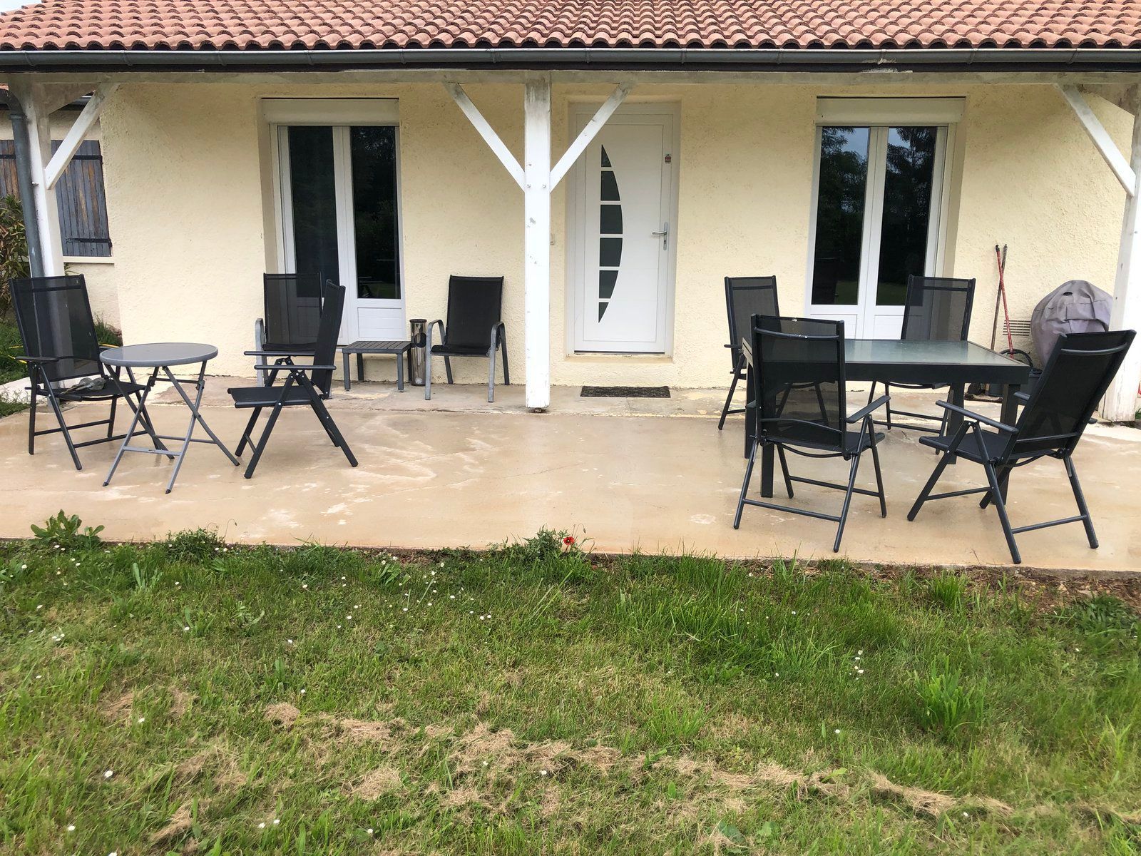 Terrasse avec chaises et tables noires à l'extérieur d'une maison couleur crème aux portes blanches et devant un premier plan gazonné
