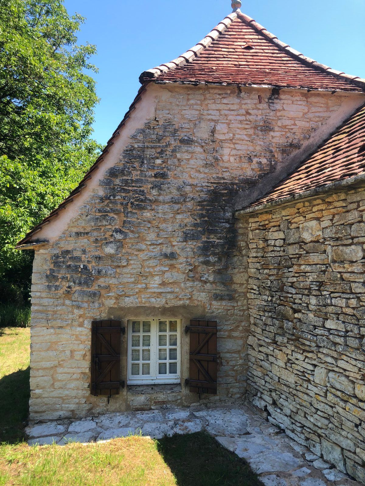 Vieille maison en pierre au toit de tuiles rouges et à petite fenêtre, donnant sur une cour ensoleillée et herbeuse