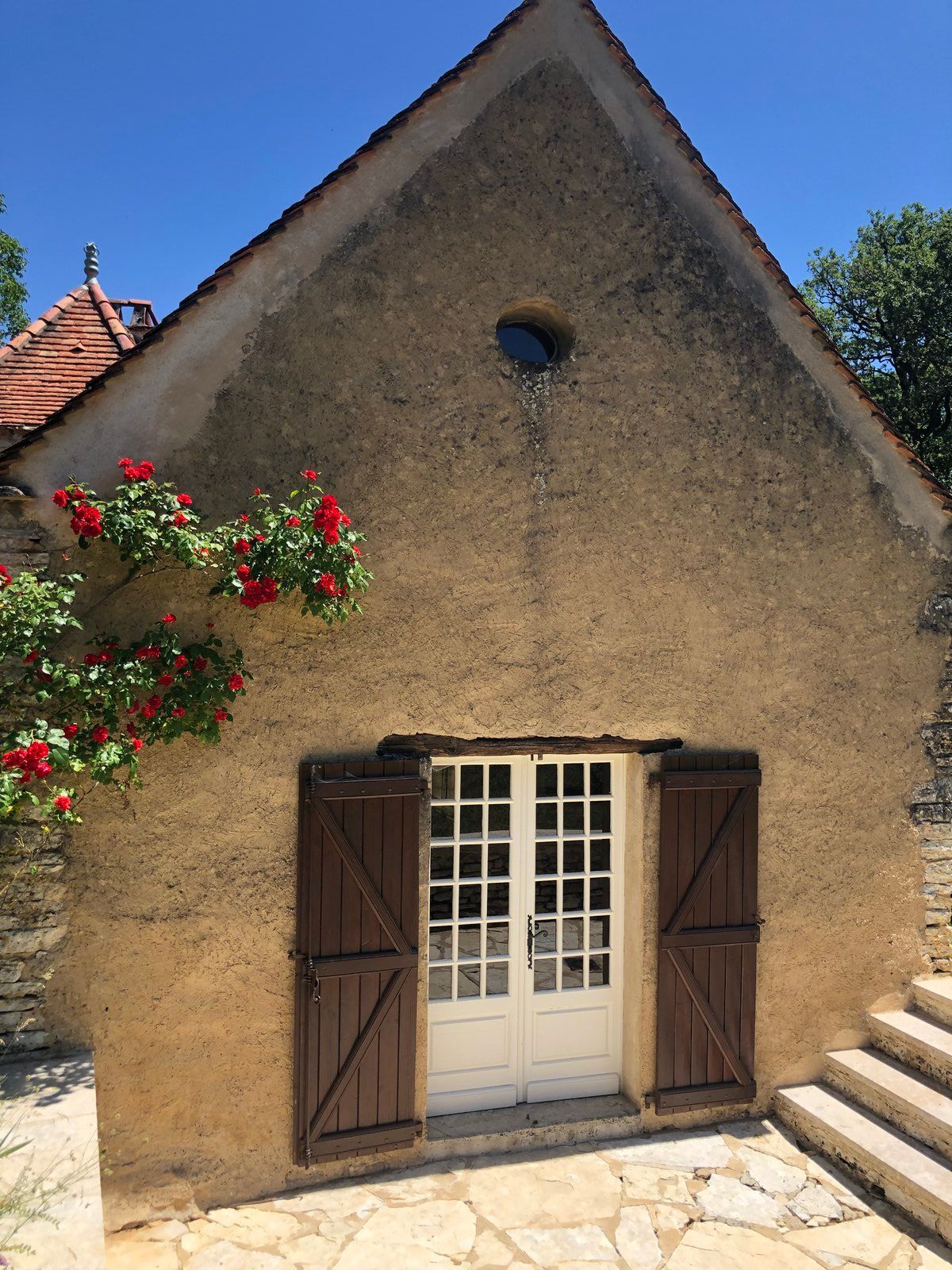Petite maison en pierre avec portes-fenêtres blanches, volets marron, fleurs rouges et marches extérieures sous un ciel bleu