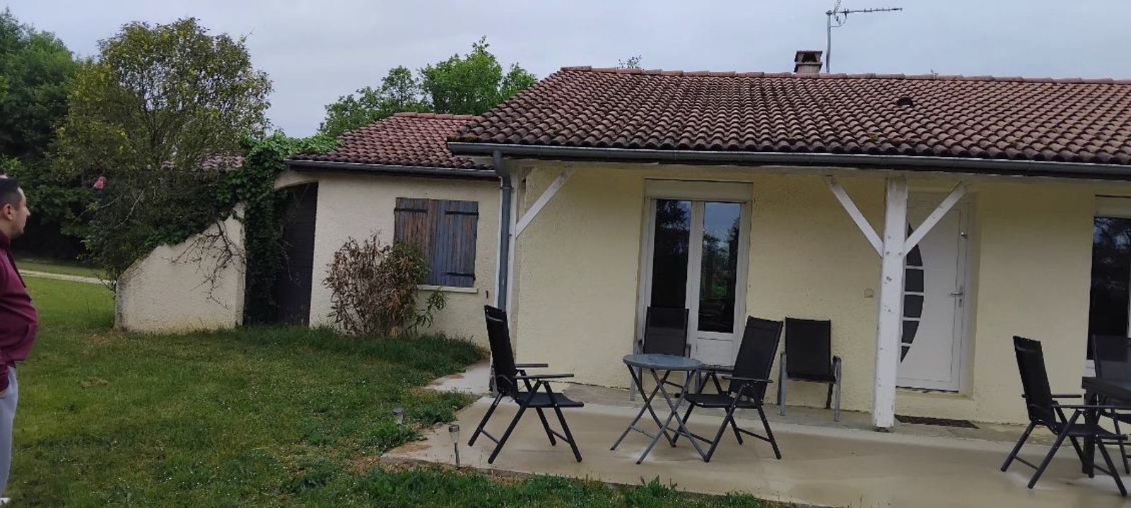 Petite maison couleur crème avec toit de tuiles, chaises et tables de patio, et une cour gazonnée sous un ciel nuageux.