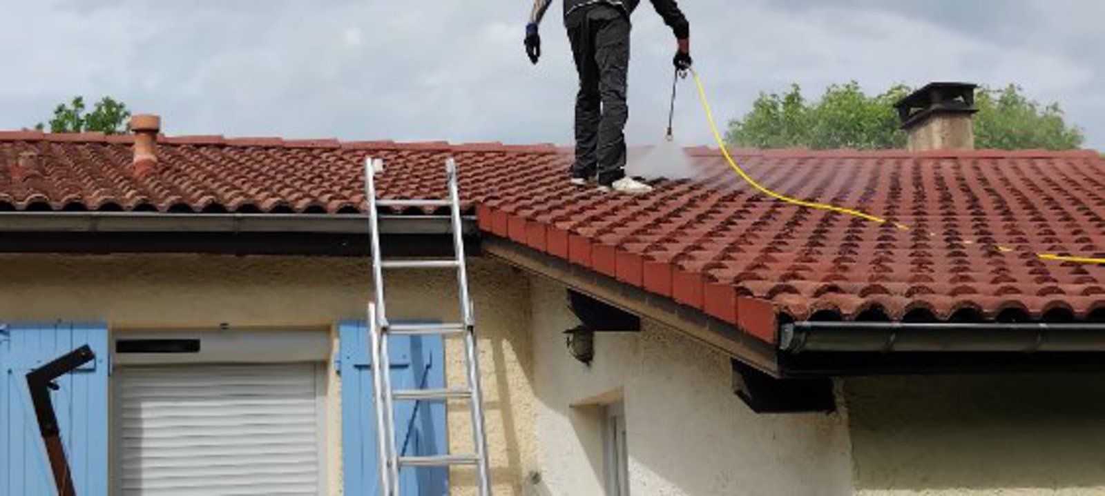 Un ouvrier, juché sur une échelle, arrose un toit de tuiles rouges avec un tuyau d'arrosage.