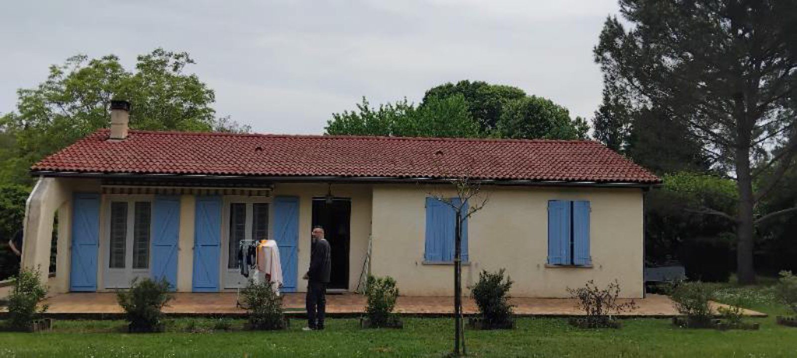 Petite maison couleur crème avec un toit de tuiles rouges, des volets bleus et deux personnes debout devant, sur une pelouse.