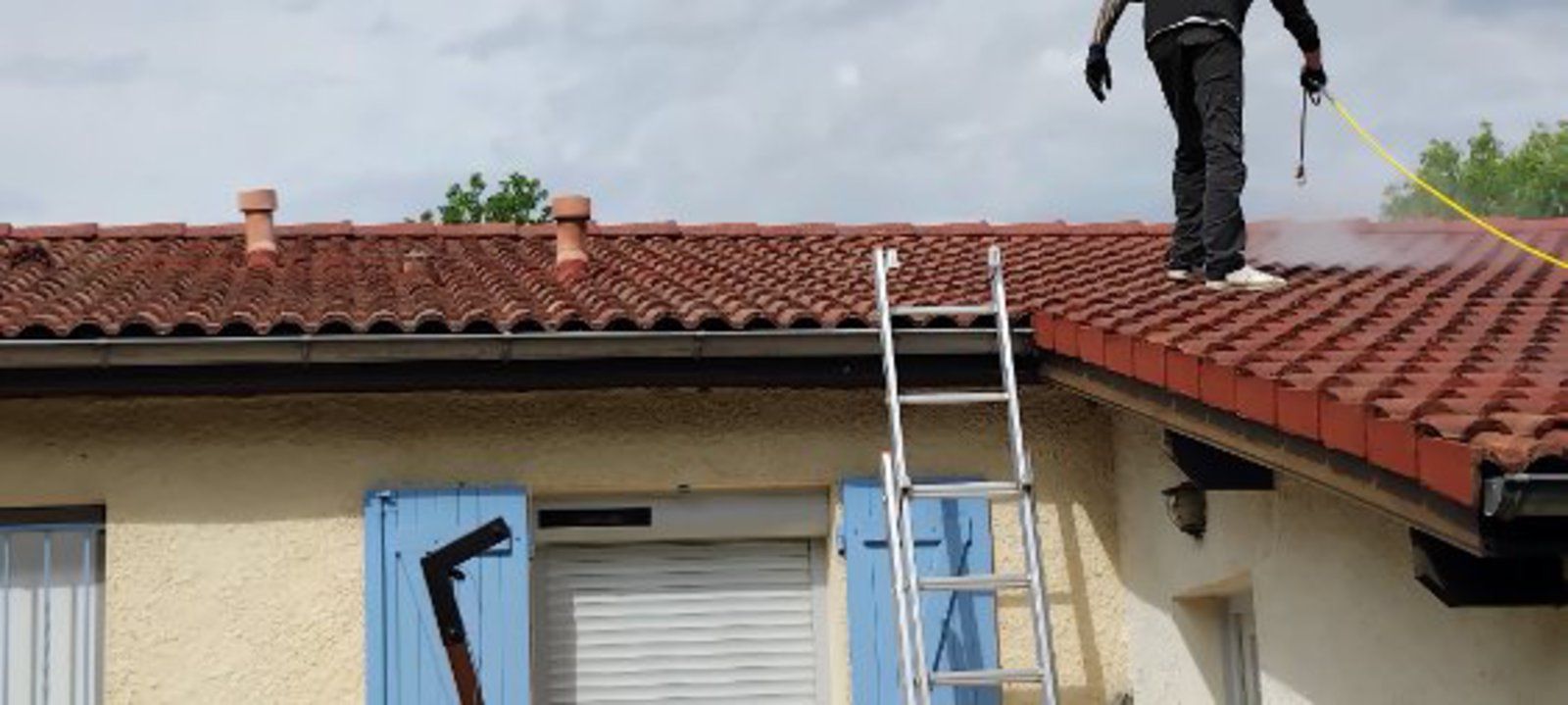 Un ouvrier, juché sur une échelle, répare un toit de tuiles rouges à côté d'une fenêtre de toit bleue.